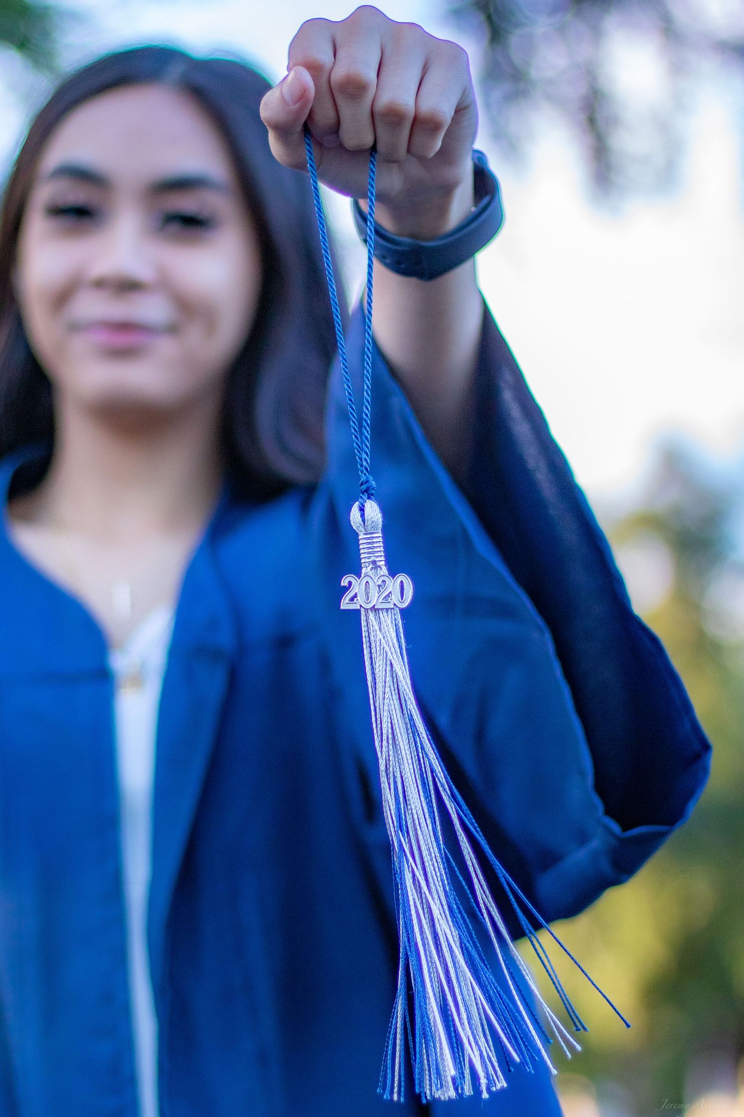 A smiling graduate in blue cap and gown holds up a 2020 graduation tassel outdoors. Stockton photographer