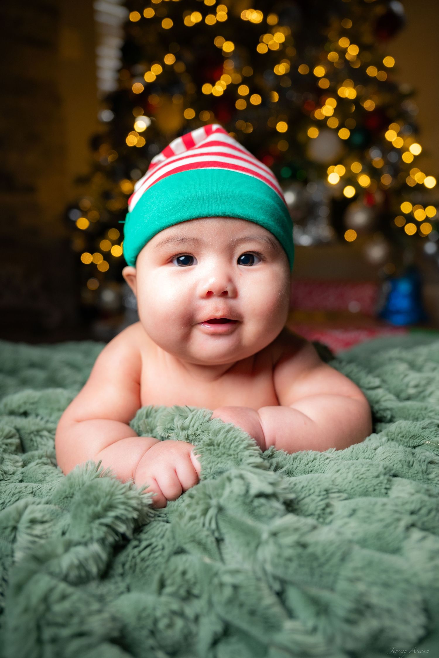 Cute baby wearing a Christmas elf hat lying on a green fuzzy blanket in front of a decorated holiday tree.
