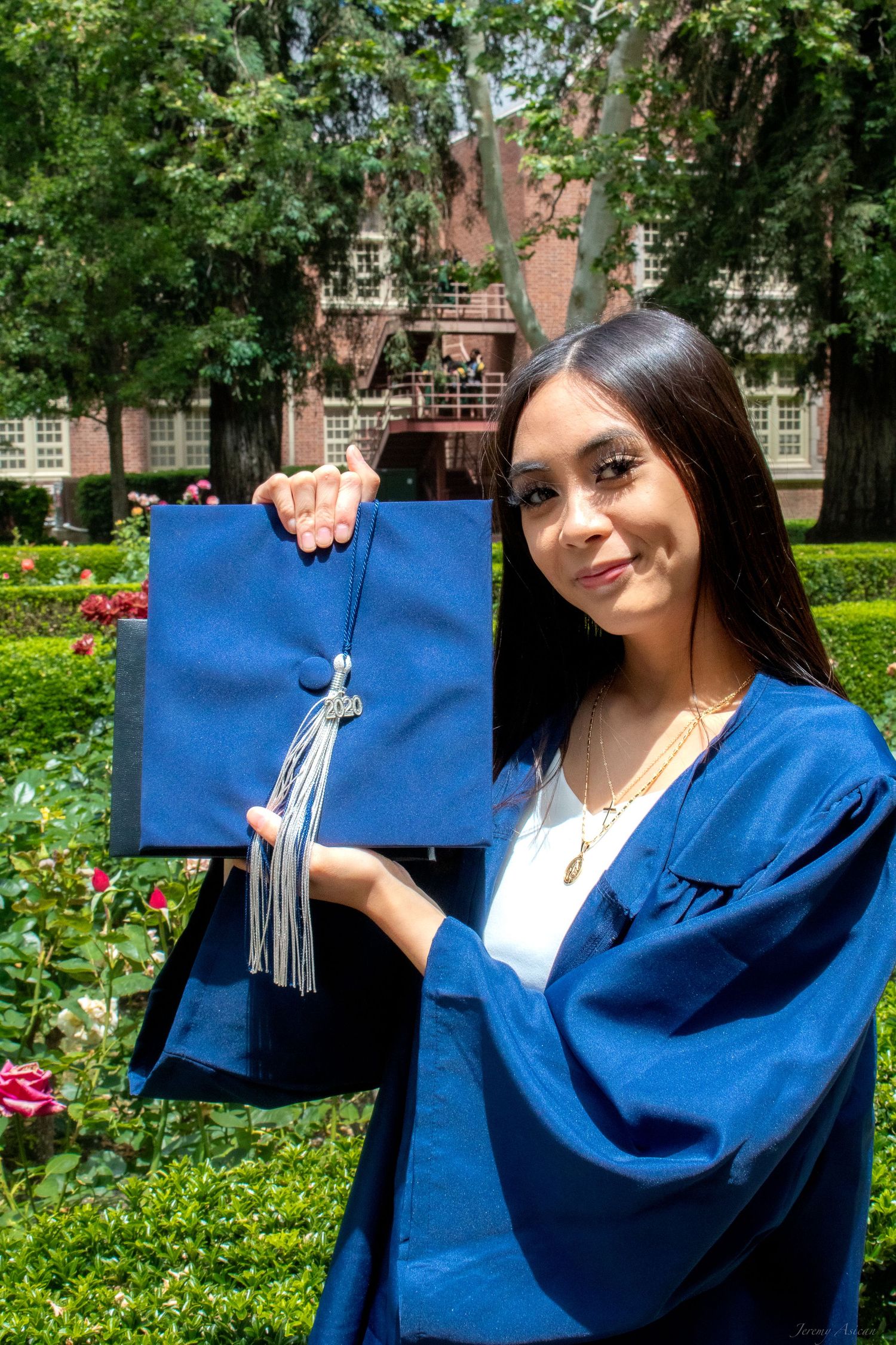 Young woman in blue graduation cap and gown holding her cap outdoors in a garden setting.