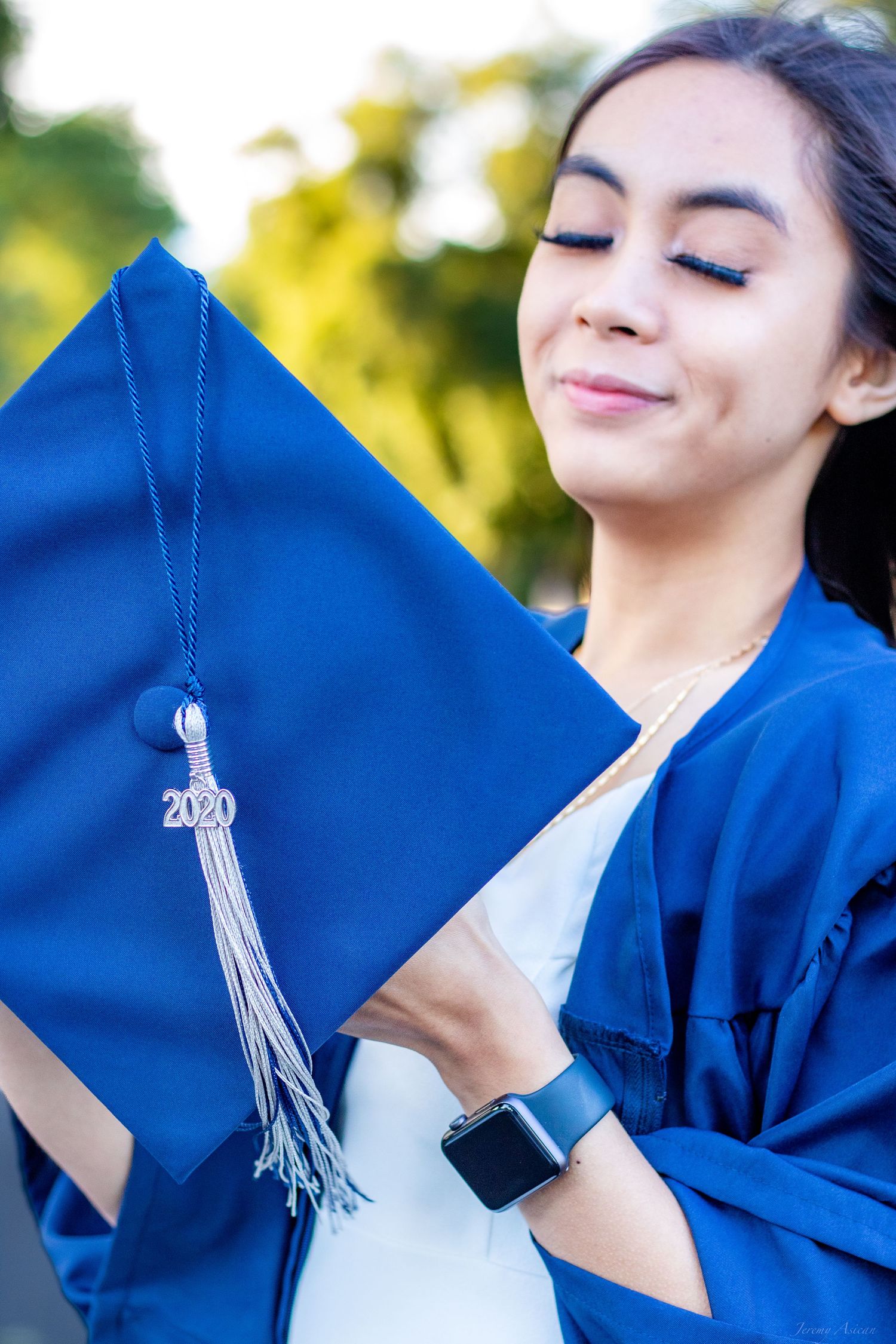 A young woman in blue graduation gown and cap with 2020 tassel smiles proudly outdoors. Stockton photographer