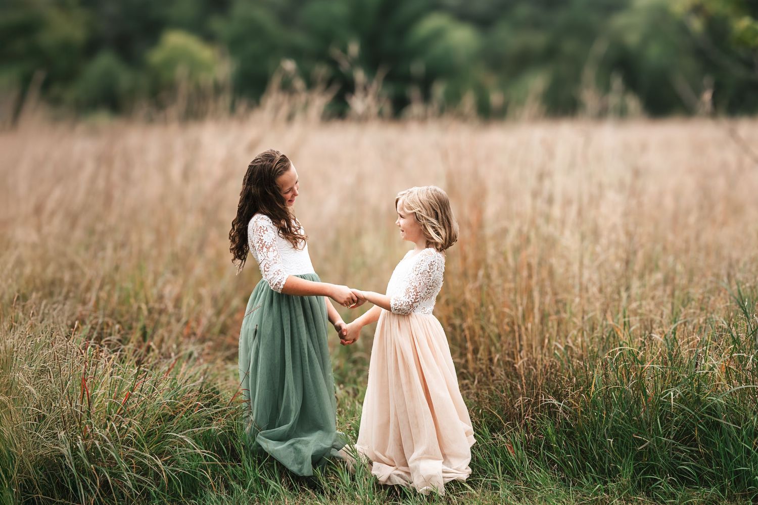 Two girls in elegant dresses holding hands in a golden field of tall grass.