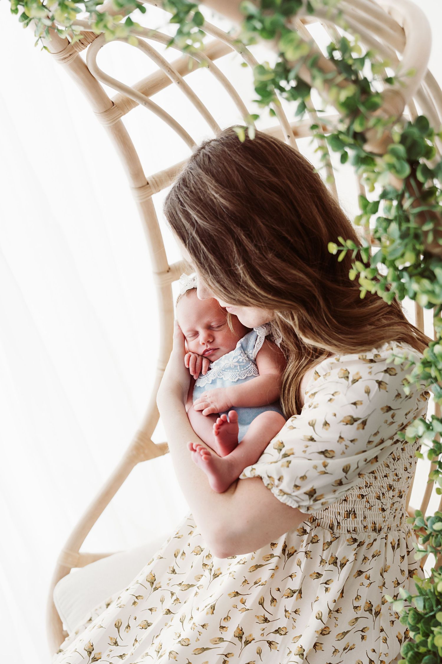 Mother sitting in a rattan swing chair kissing her newborn baby in a bright studio setting.