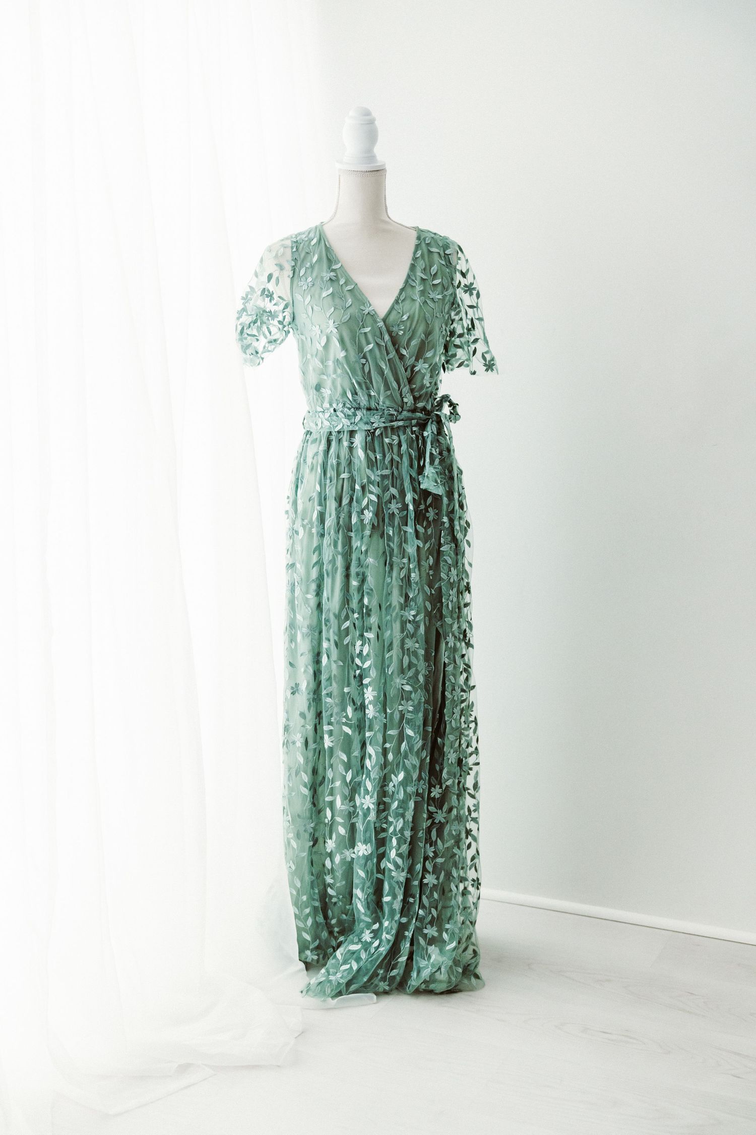 Sage green leaf lace wrap maternity dress with tie waist on mannequin.