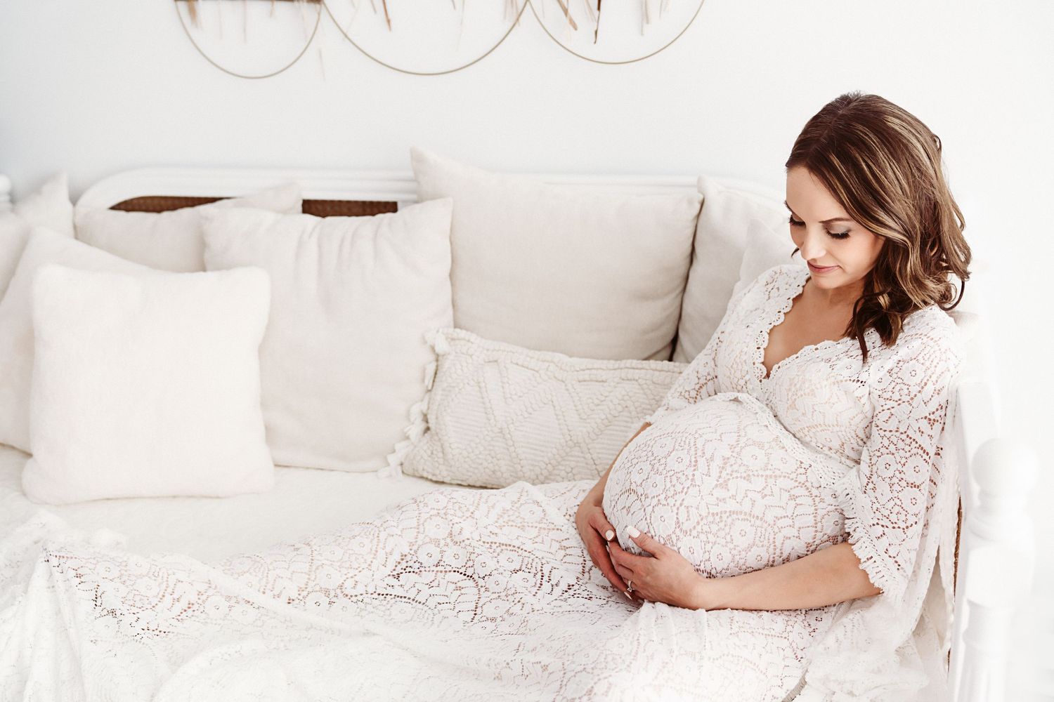 A pregnant woman in a white lace dress sits on a white bed, lovingly gazing at her baby bump.