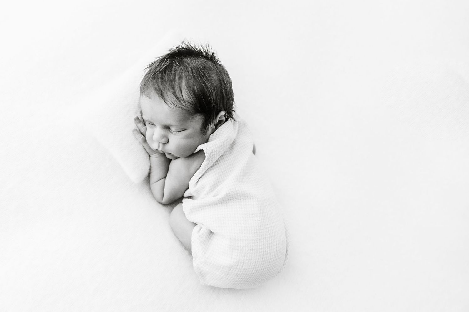 Black and white photo of curled sleeping newborn baby on white backdrop.