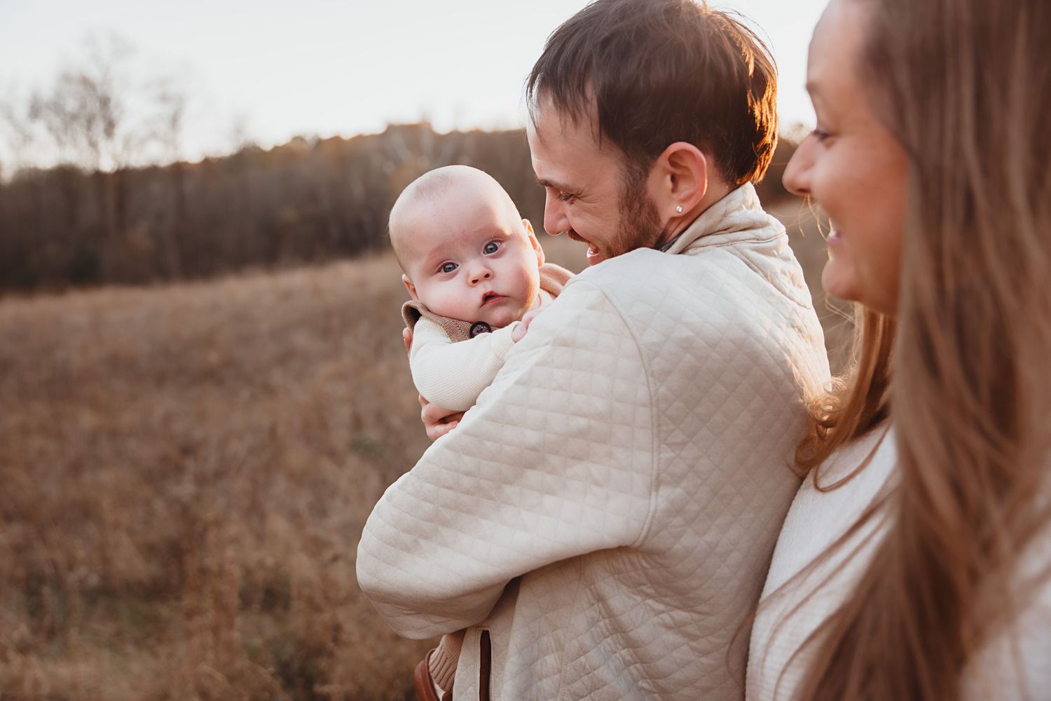 Father holds baby close while mother smiles in a golden autumn field.