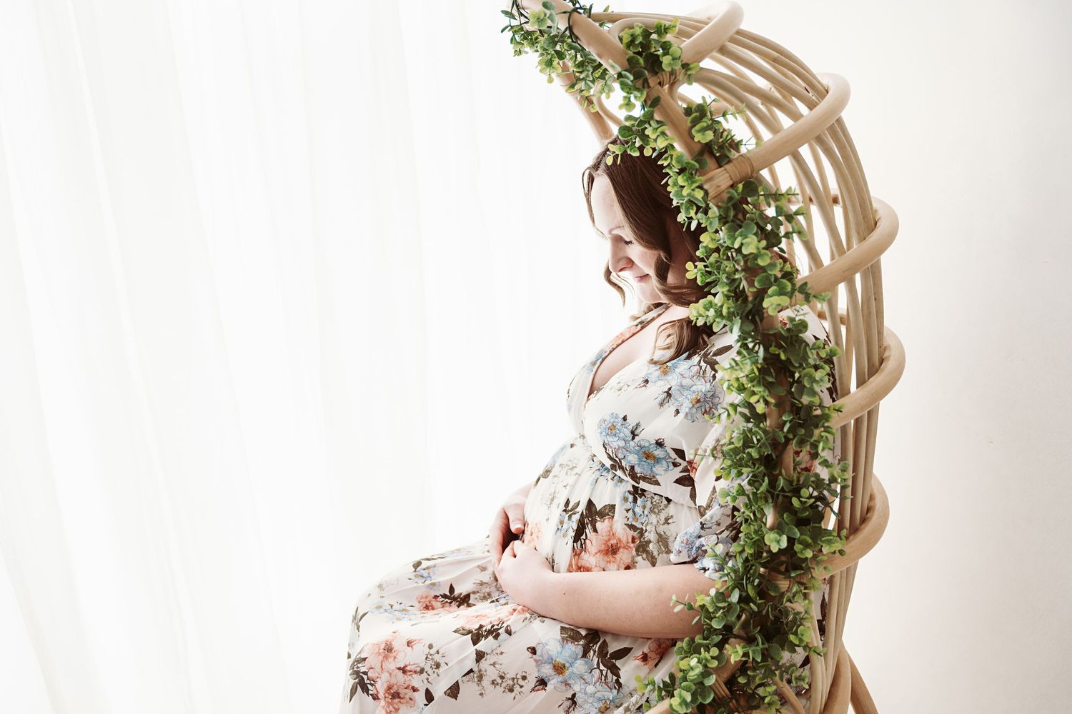 Pregnant woman in floral dress sitting in rattan chair adorned with greenery garland, smiling softly.