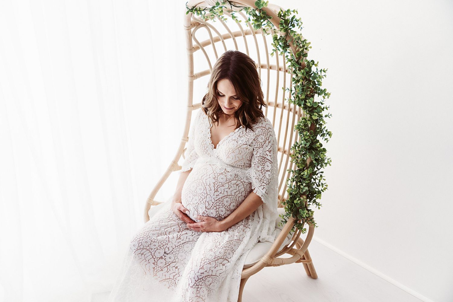 Pregnant woman in white lace dress sitting on rattan swing chair decorated with greenery.