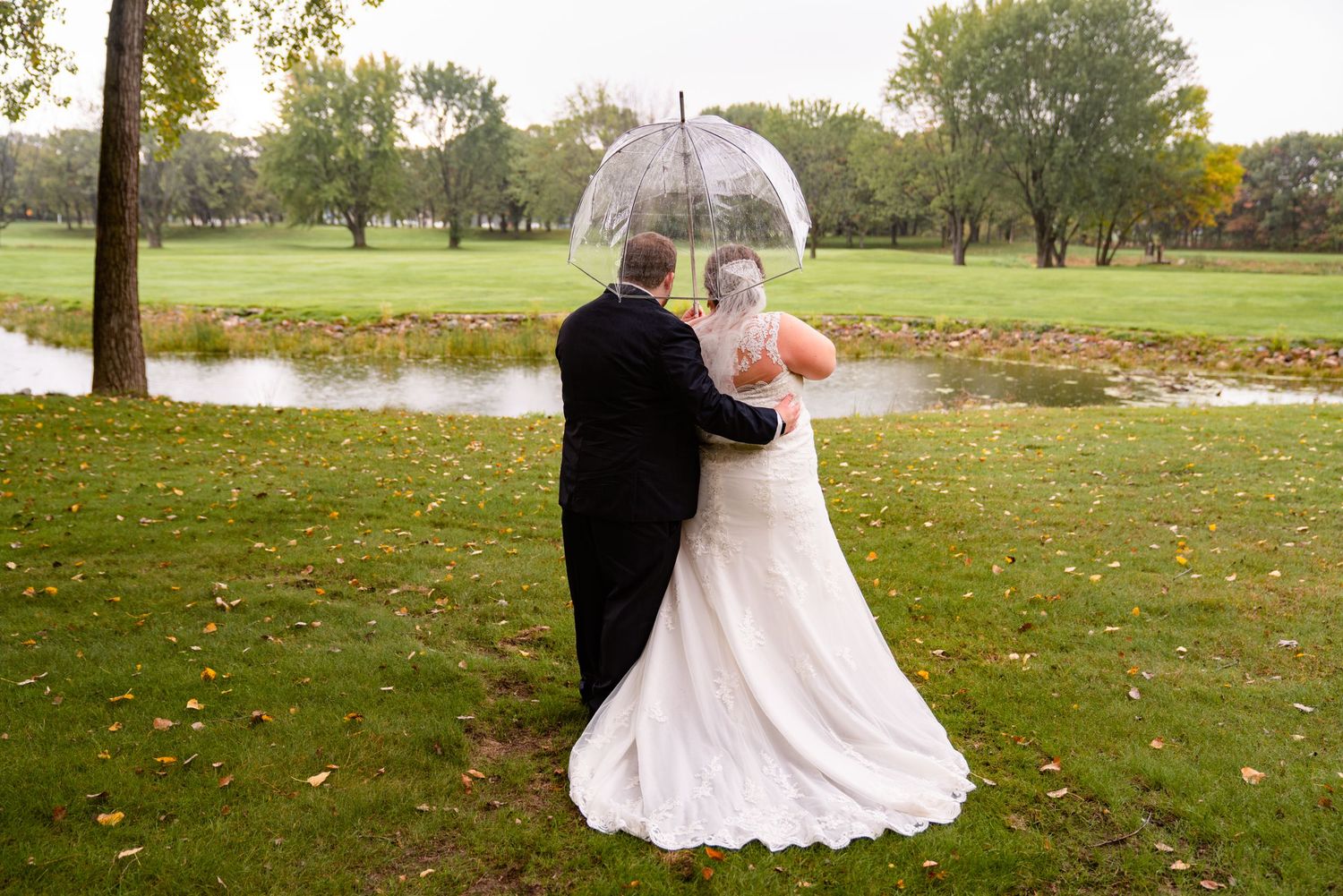 Bride and groom sharing romantic moment under clear umbrella on rainy wedding day by a pond.