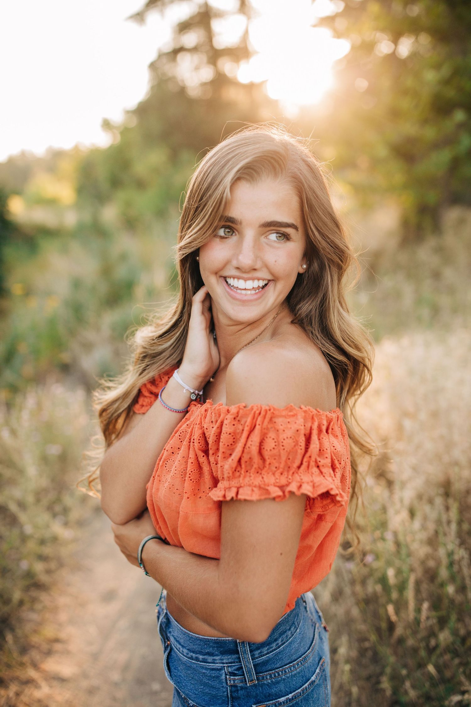Smiling young woman in orange off-shoulder top and jeans poses on a sunlit nature trail.