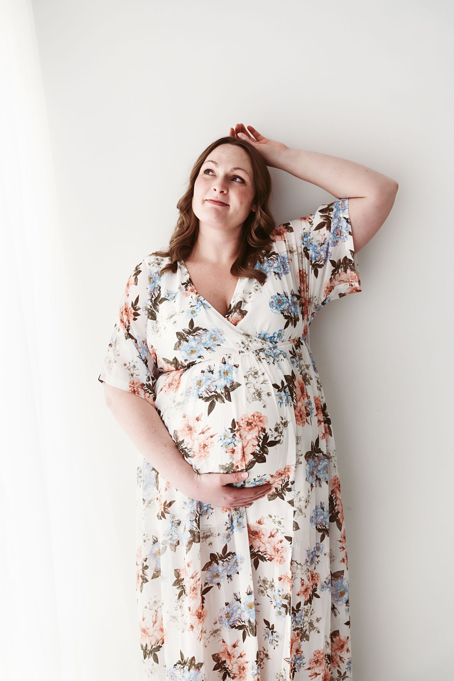 Pregnant woman in floral wrap dress leaning against white wall, cradling her baby bump.