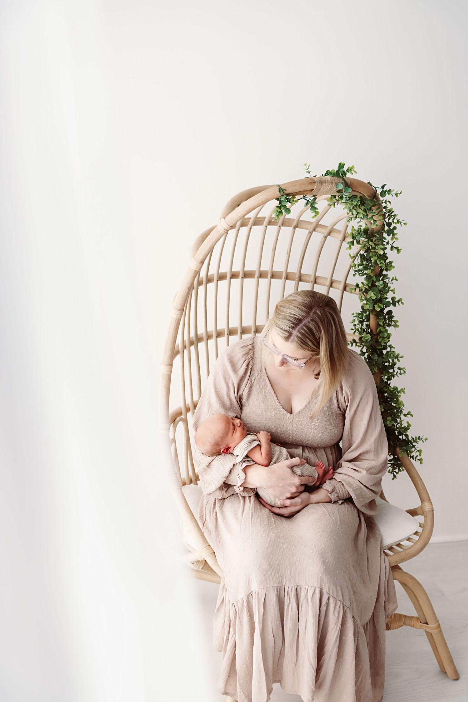 Mother in blush dress cradles newborn in rattan hanging chair with greenery.