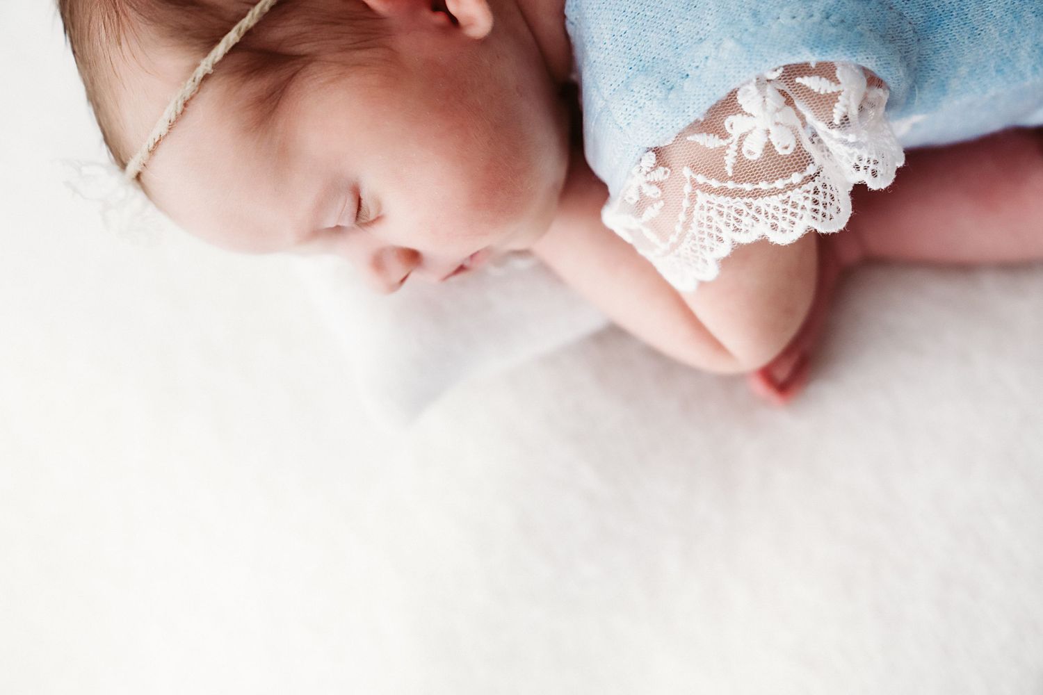 A sleeping newborn baby wearing a blue lace outfit and delicate headband rests on a soft white blanket.