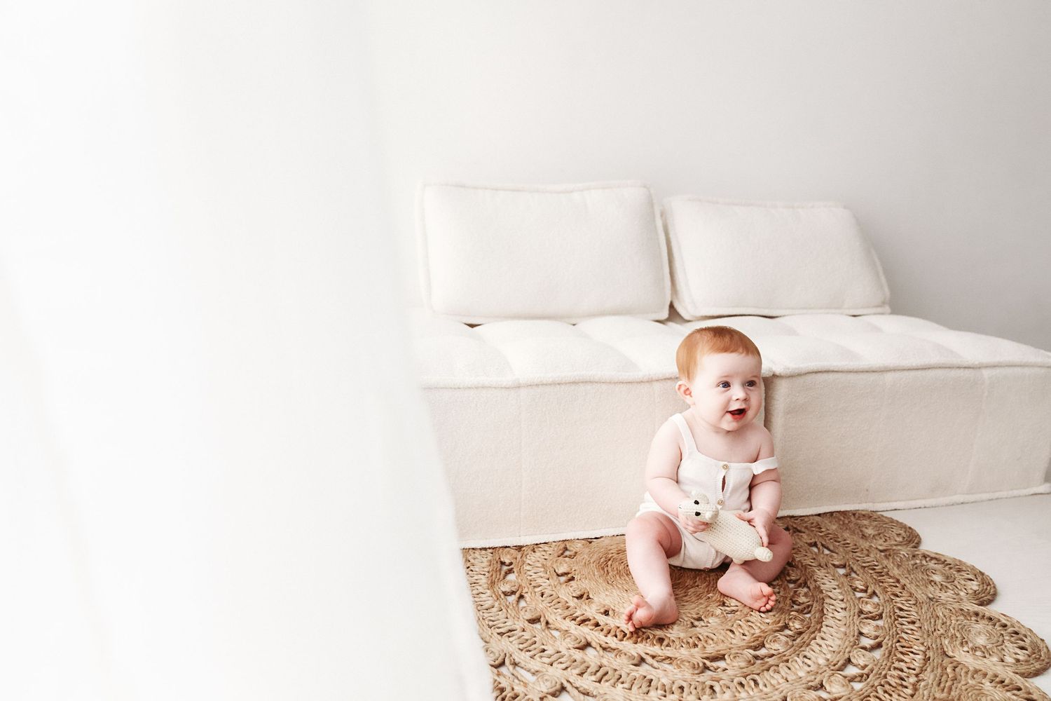 A cheerful red-haired baby sits on a jute rug holding a stuffed toy near a white sofa.