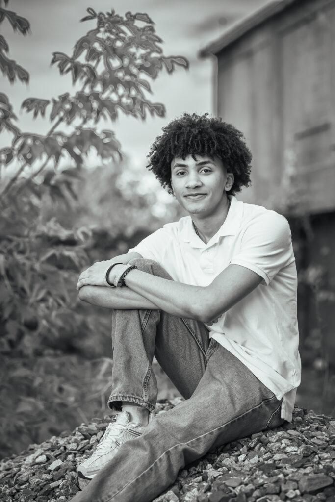Black and white portrait of a young man with curly hair sitting on rocks, wearing a white polo shirt and jeans.