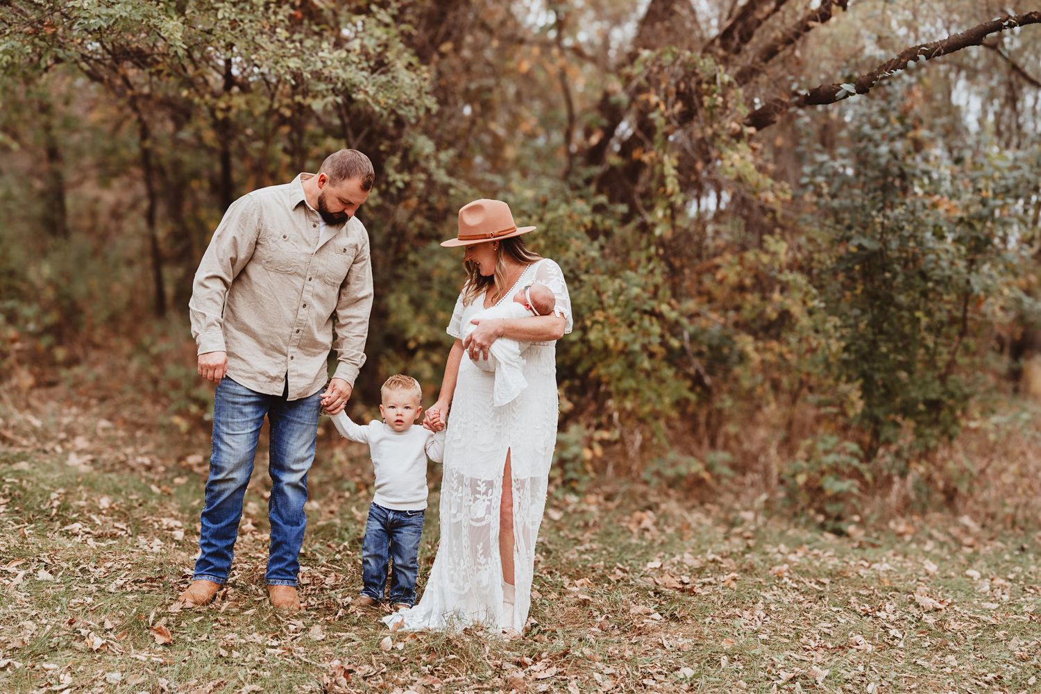 Young family with toddler and newborn baby walking together on a leaf-covered autumn forest path.