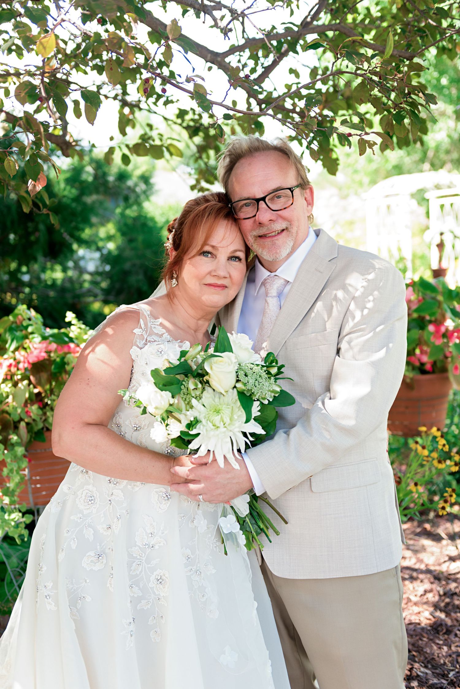 Smiling bride and groom pose together in garden holding white floral bouquet.
