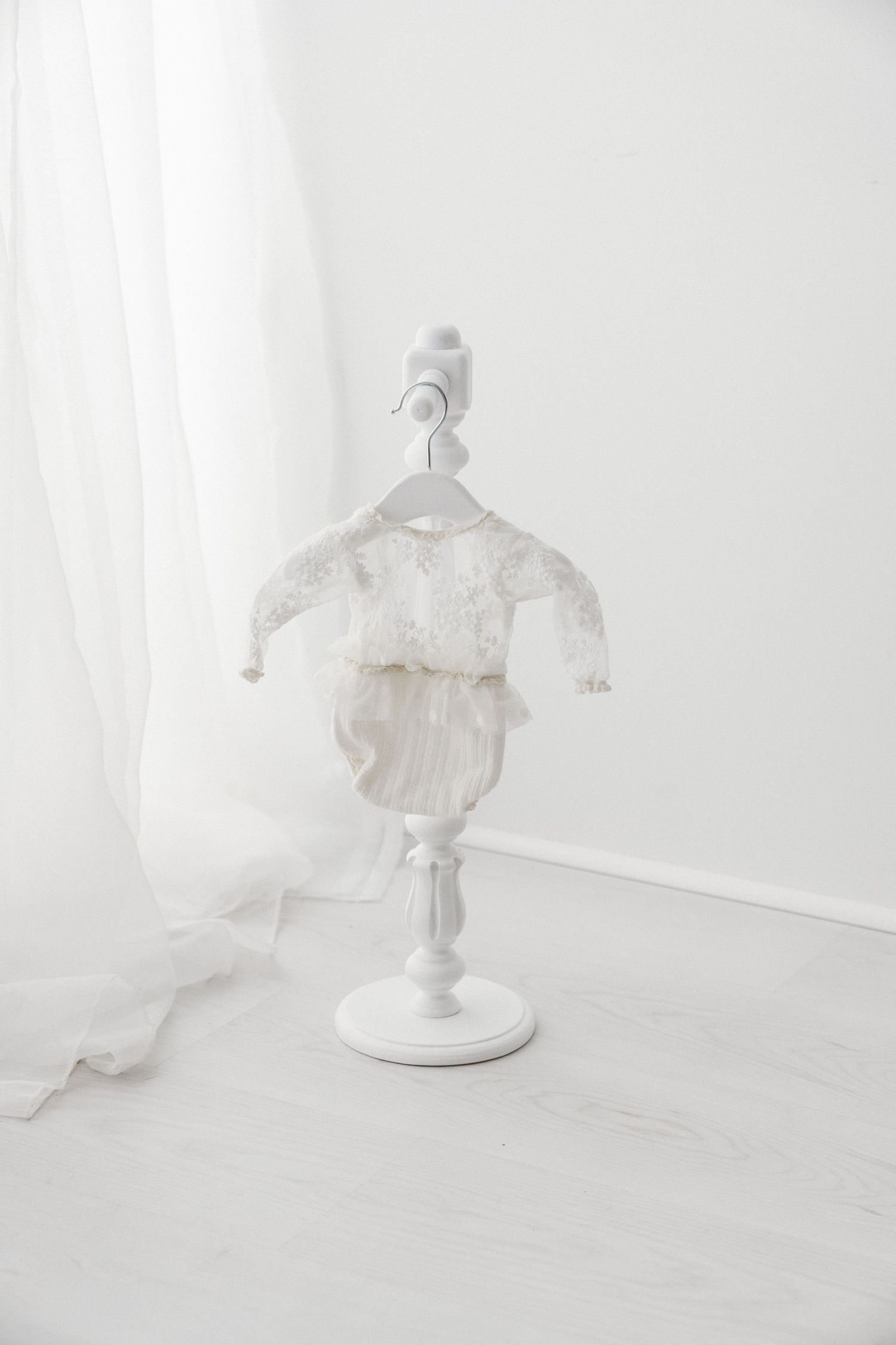 White lace baby romper with tulle skirt hanging on a decorative white mini stand in a bright minimalist white room.