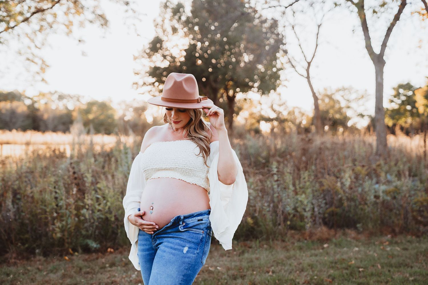 Pregnant woman in white top and jeans adjusting her brown hat while cradling her bump in a golden field.