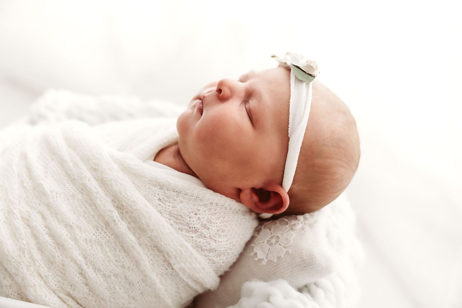 Close-up of peaceful sleeping newborn baby swaddled in white wrap wearing a delicate floral headband.
