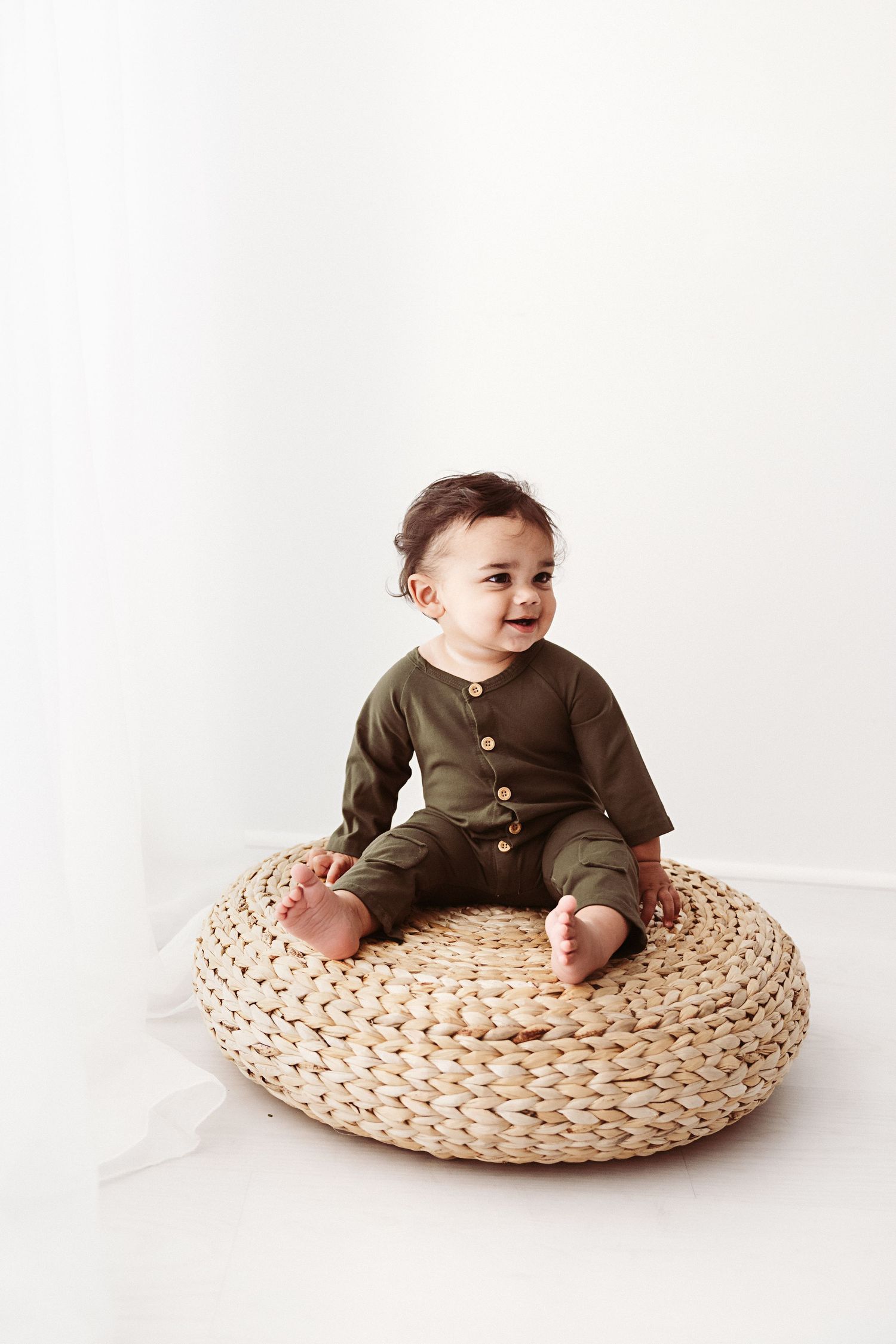Happy baby in olive green button romper sitting on woven rattan pouf, smiling.
