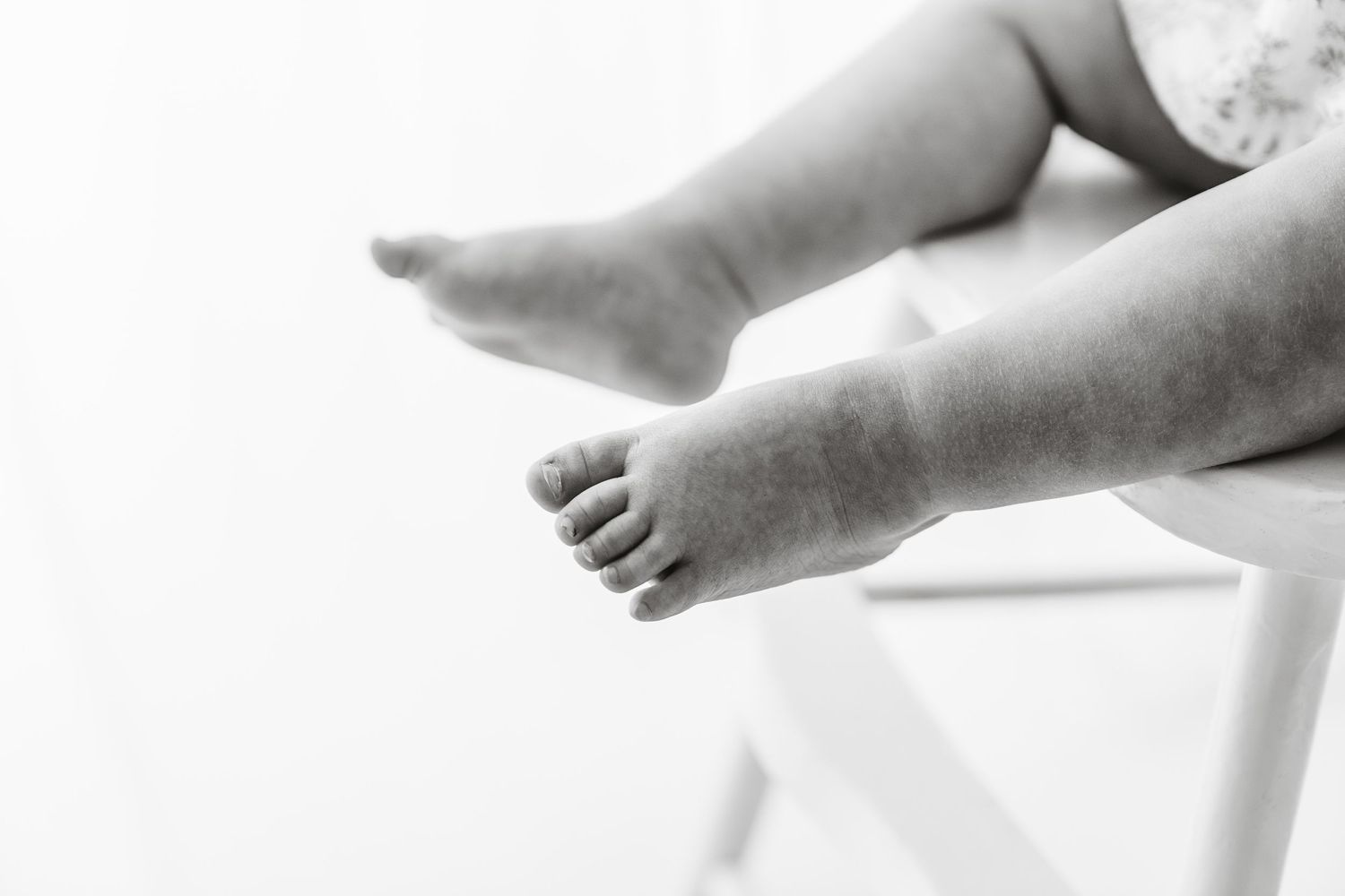 Black and white close-up of chubby baby feet dangling off the edge of a white chair.