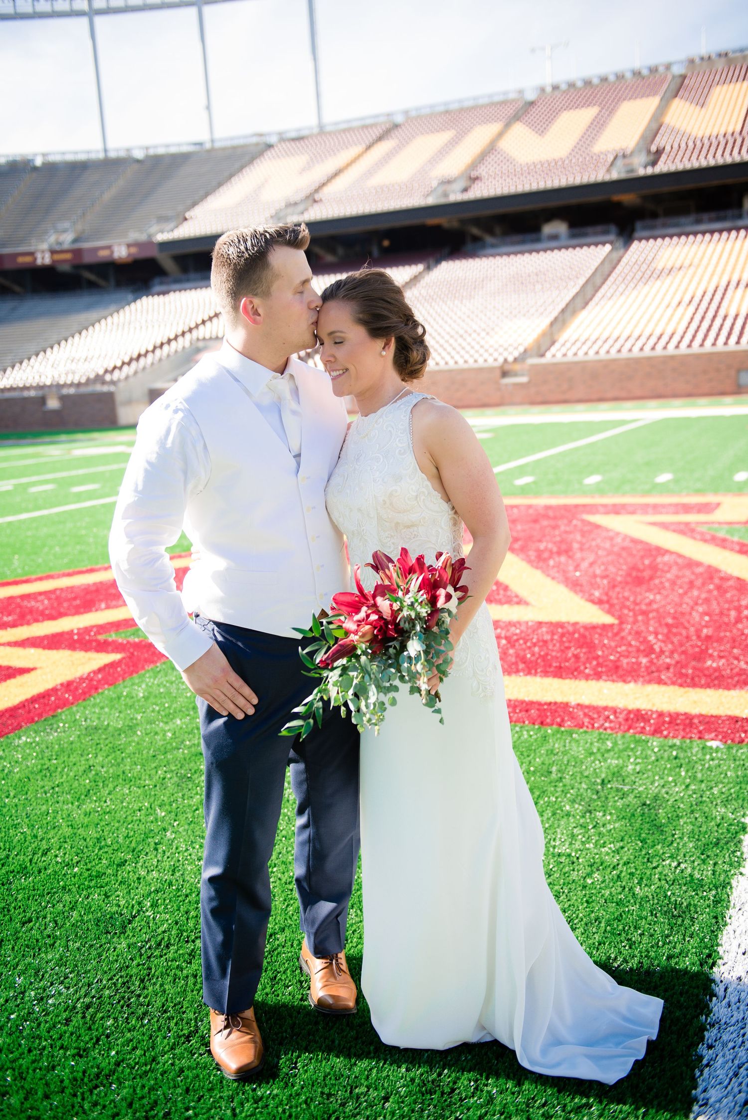Wedding couple posing romantically on a football stadium field, bride holding red and green bouquet.