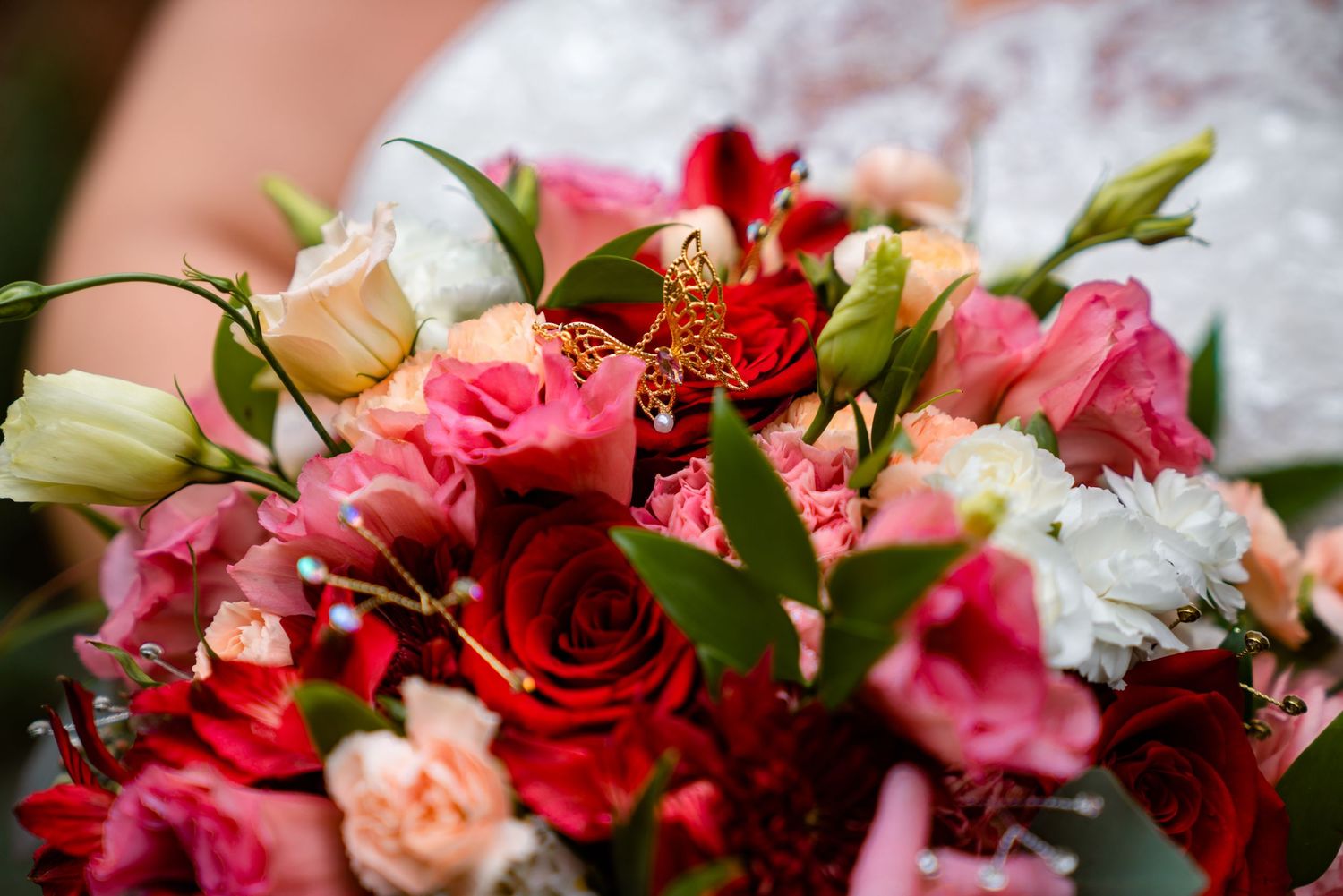 Close-up of bridal bouquet featuring red roses, pink carnations, and white flowers with gold butterfly.
