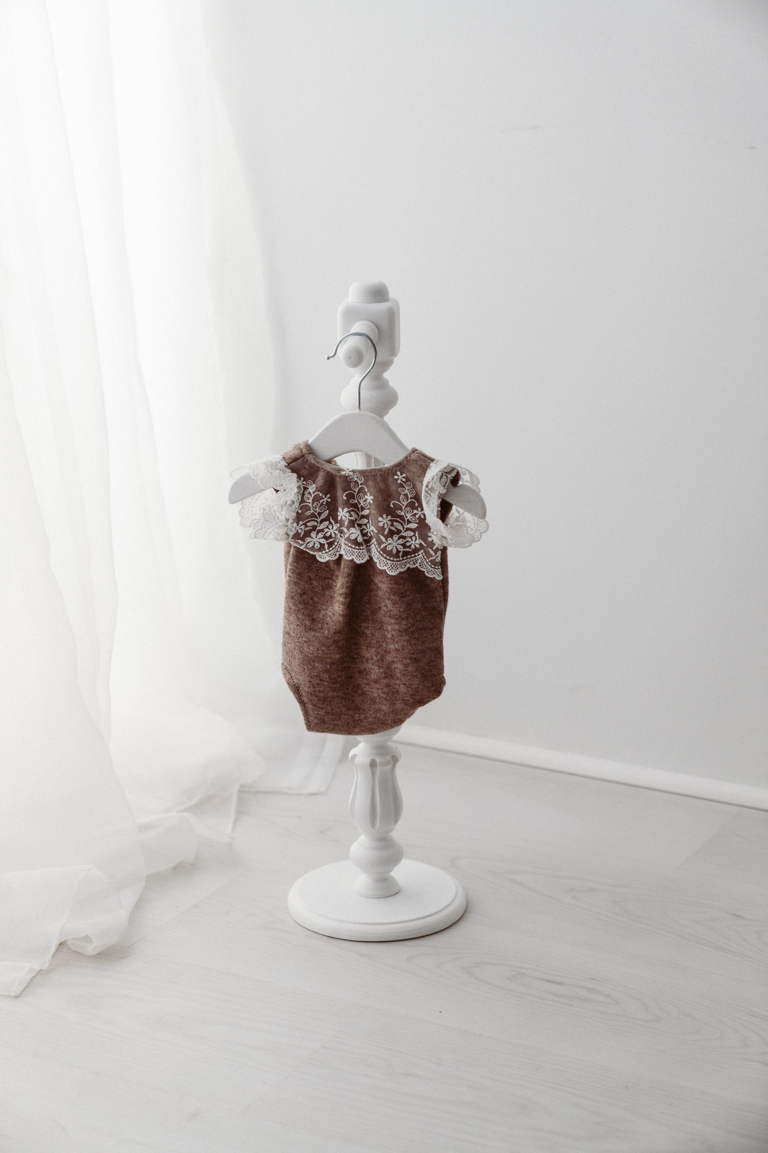 Brown knit baby romper with white lace collar detail hanging on a white decorative mini stand.
