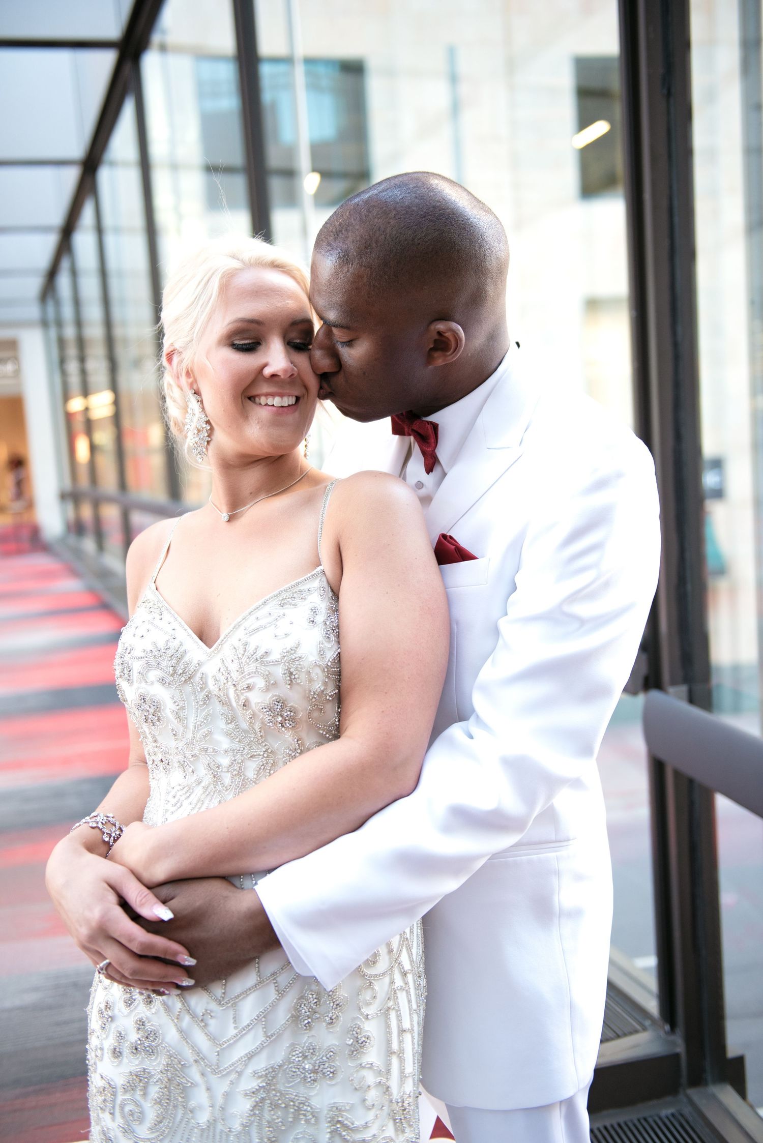 Groom in white tuxedo tenderly kissing bride on cheek while embracing her outside glass building.