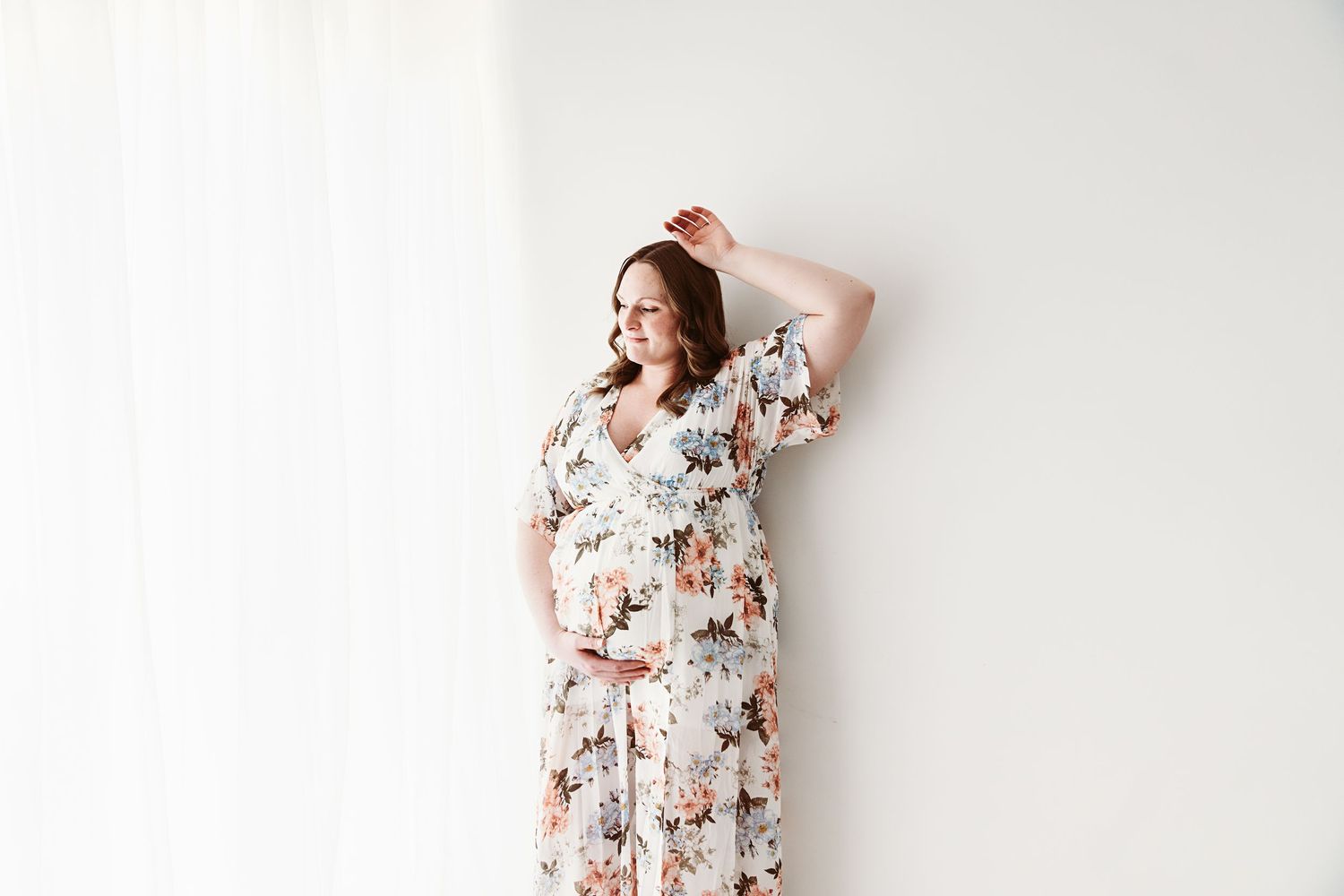 Pregnant woman in floral dress leaning against white wall, gently touching her baby bump.