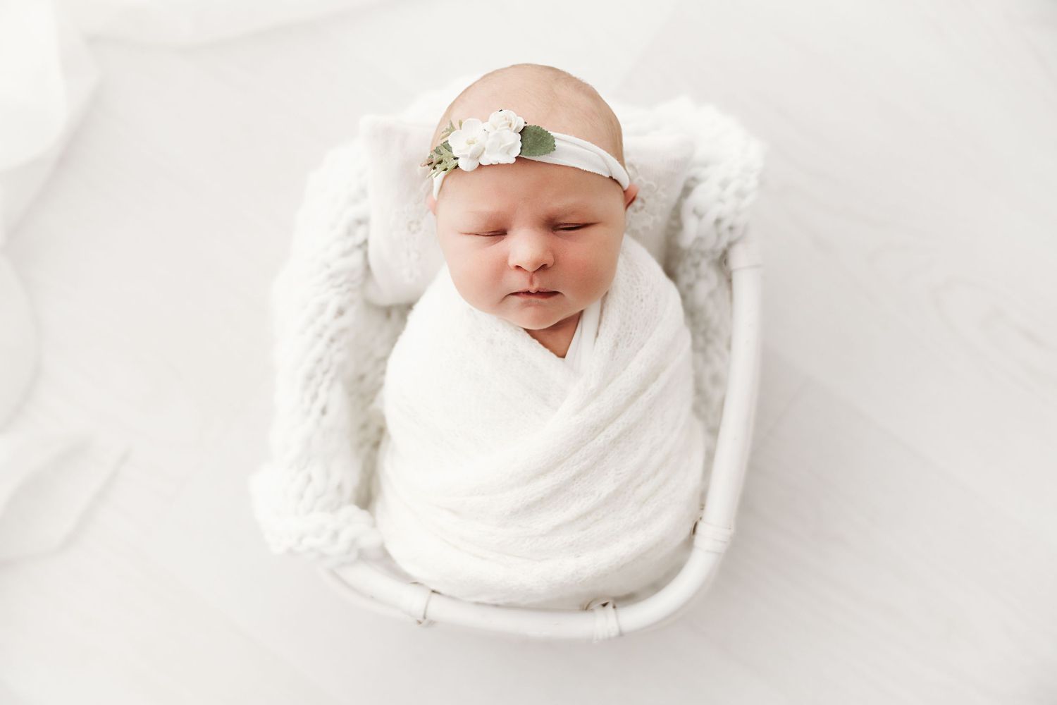 Newborn baby wrapped in white swaddle sleeping peacefully in a small white basket.