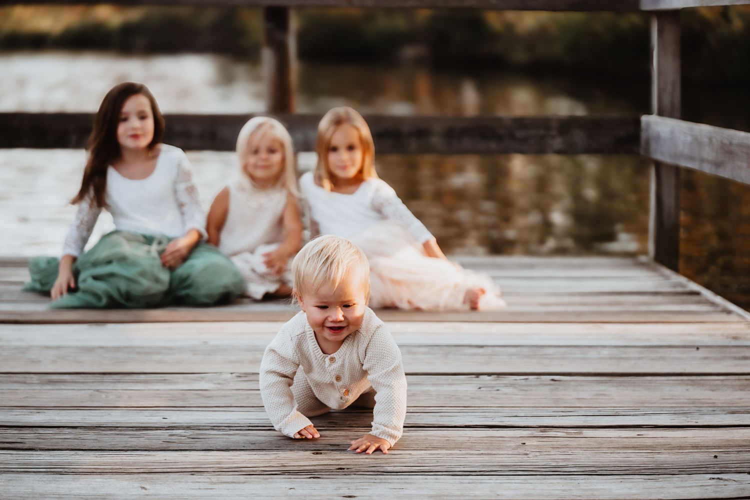 Toddler crawling on wooden dock while three girls sit together in background.
