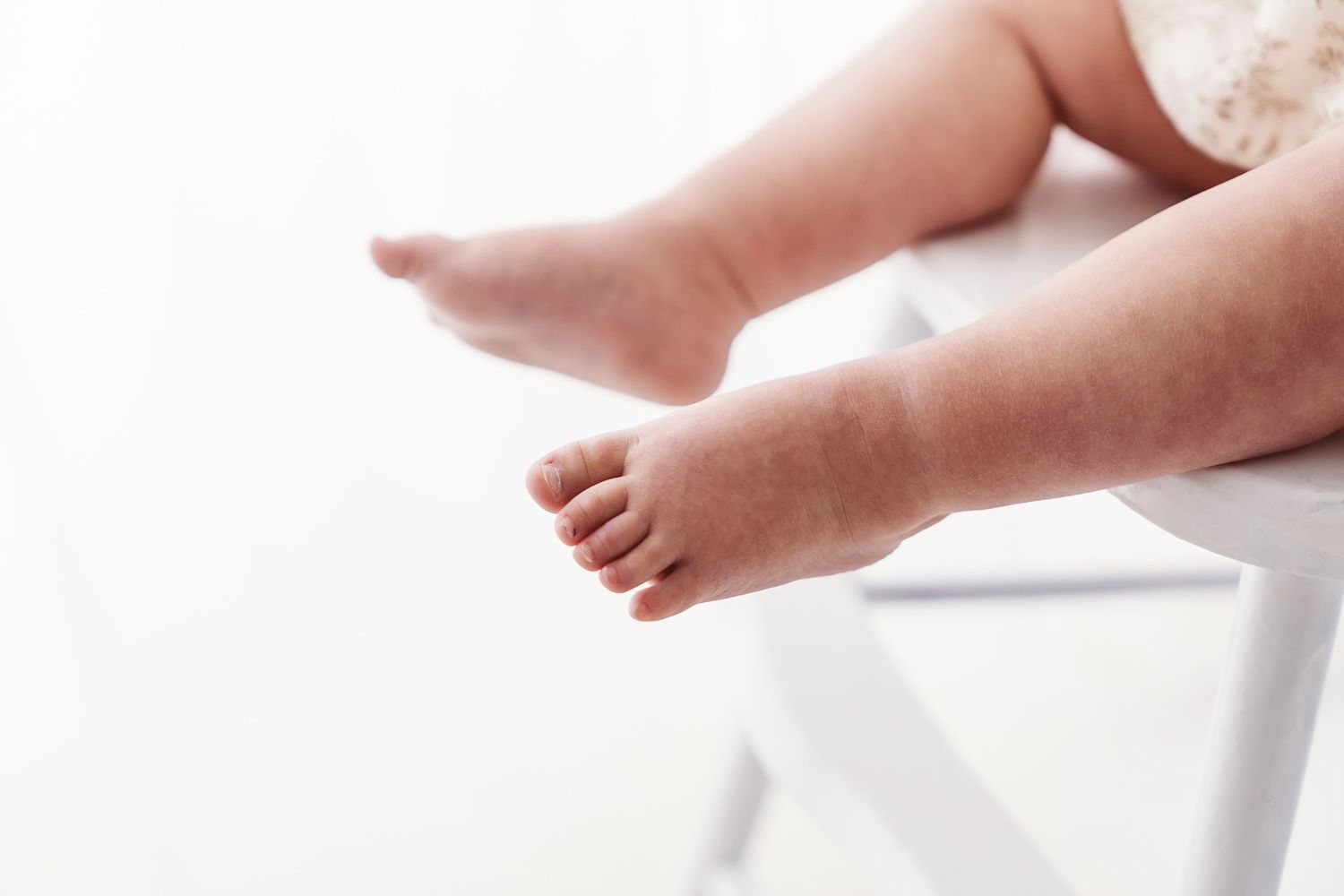 Close-up of a baby's chubby feet dangling off the edge of a white chair on a white background.