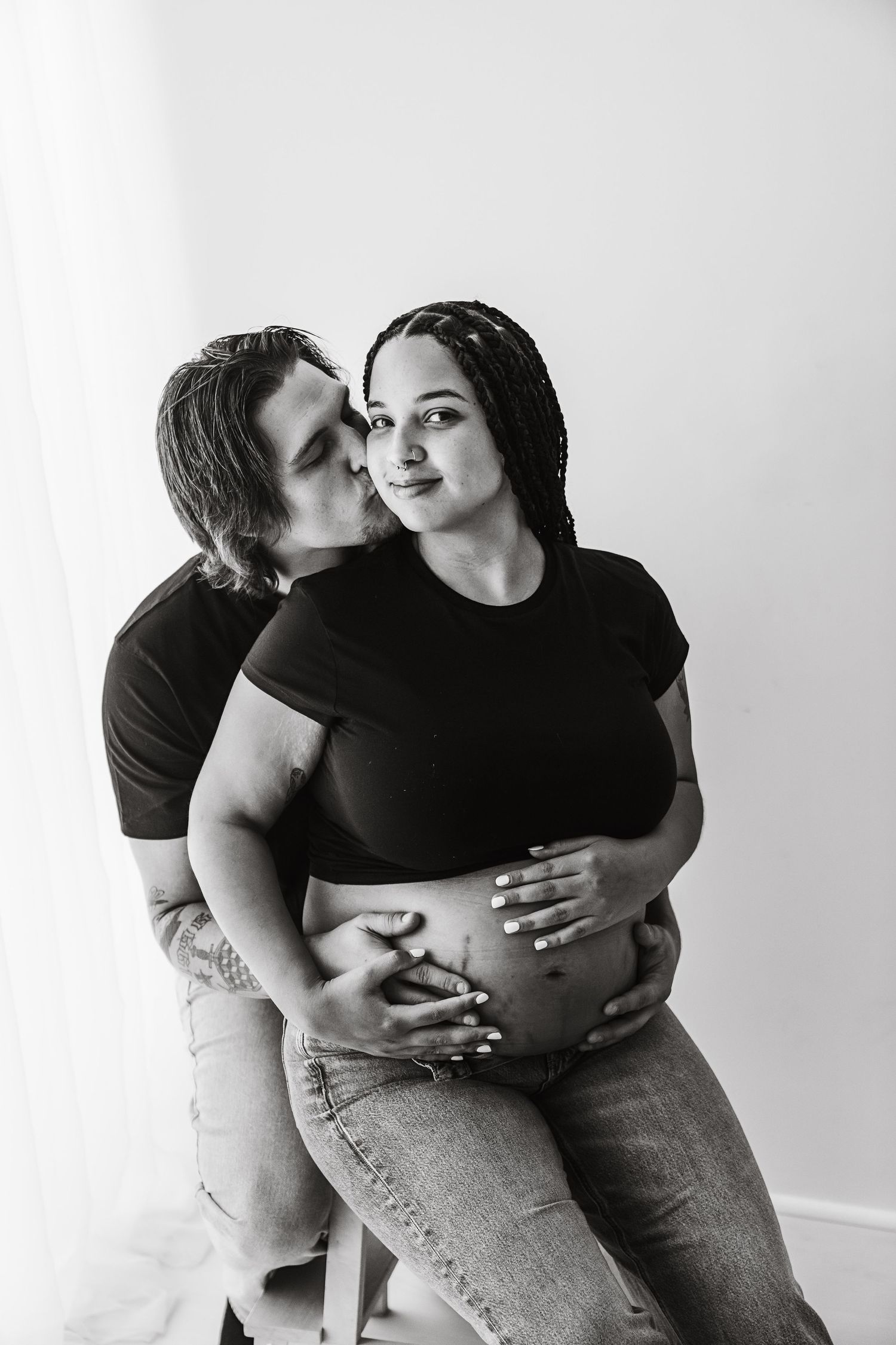 Black and white photo of a couple, man kissing pregnant womans cheek while holding her belly.