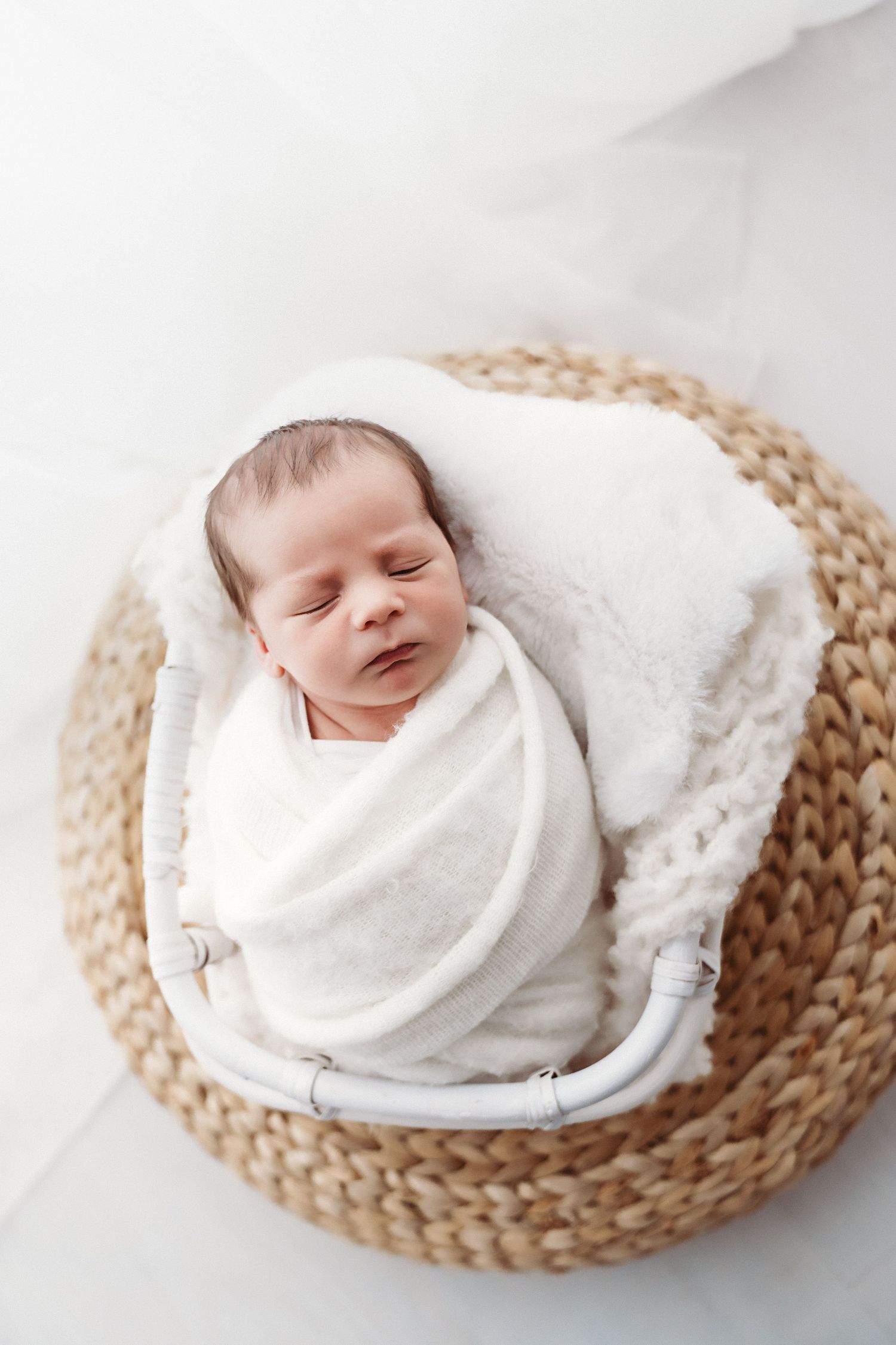 A swaddled newborn baby sleeps peacefully in a small white basket on a woven round mat.
