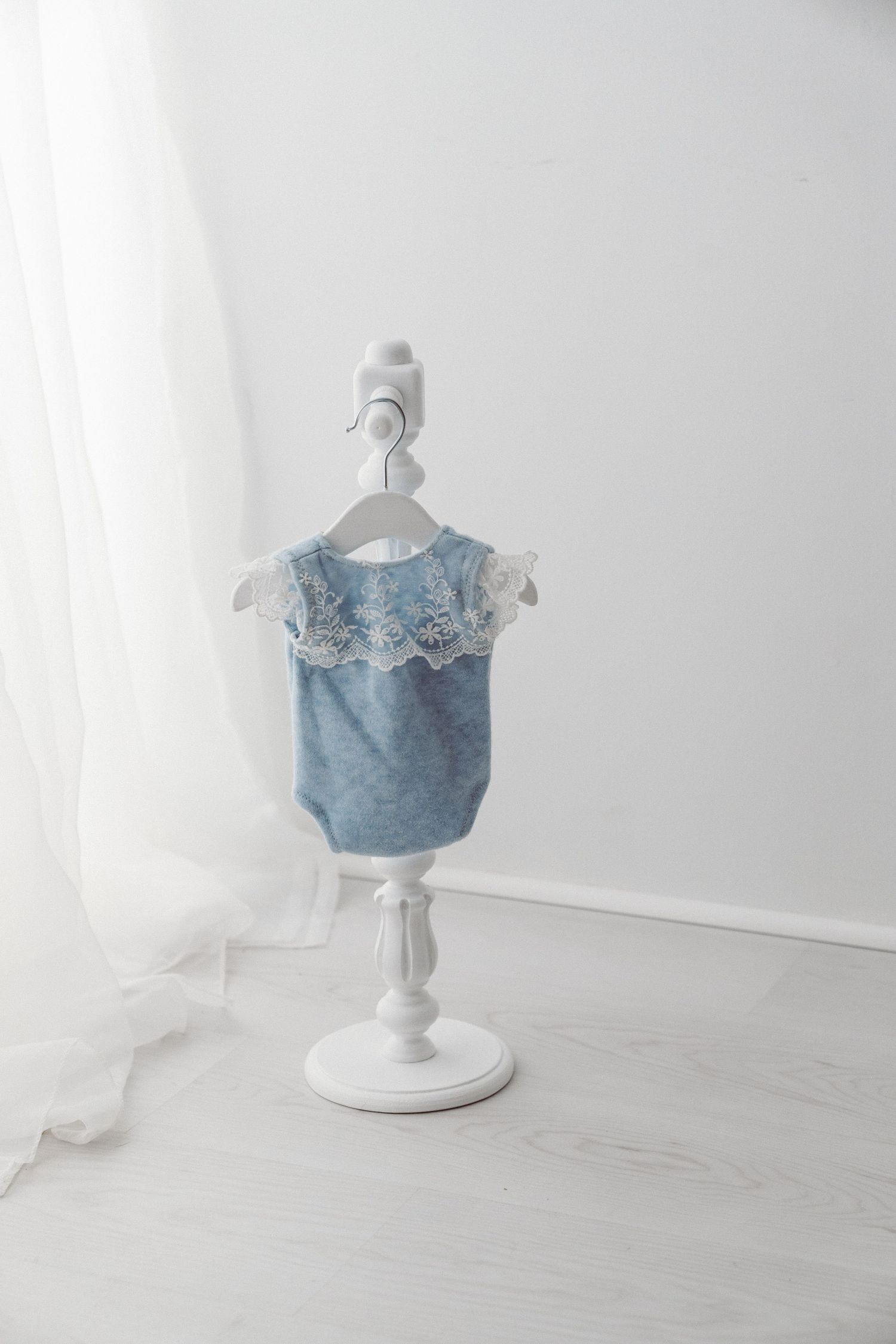 Blue denim baby romper with white lace collar displayed on a white decorative mini clothing stand.