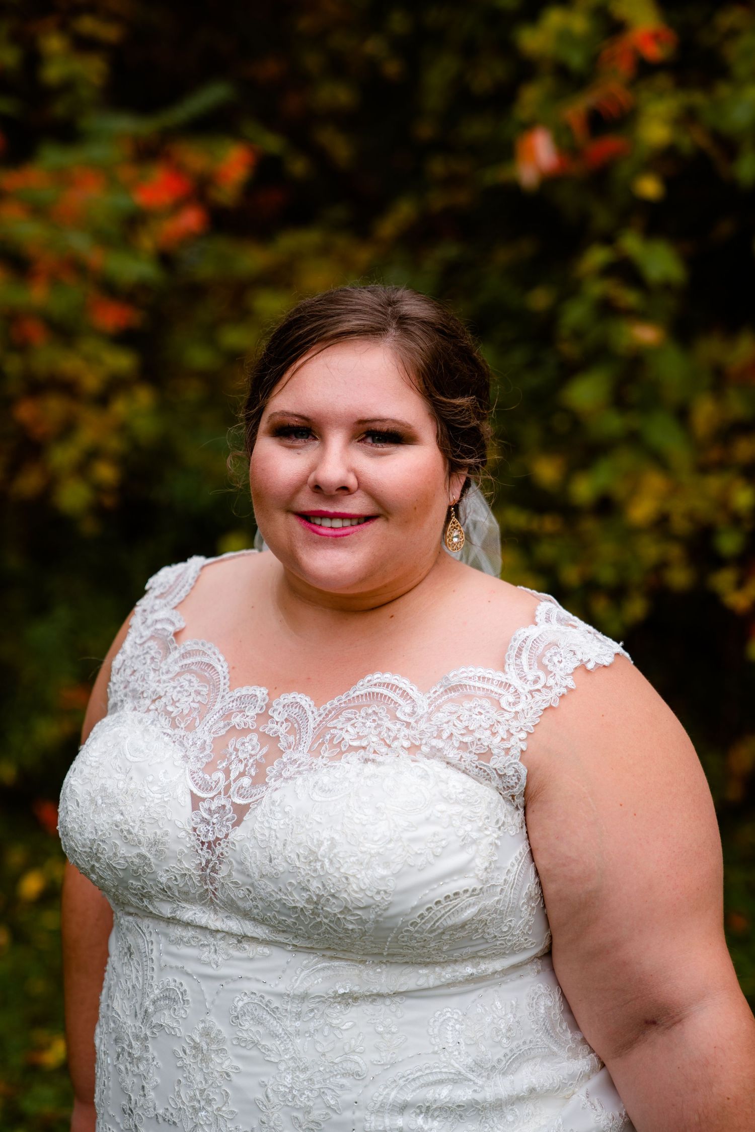 Plus-size bride smiles warmly in lace cap-sleeve wedding gown against vibrant fall foliage background.