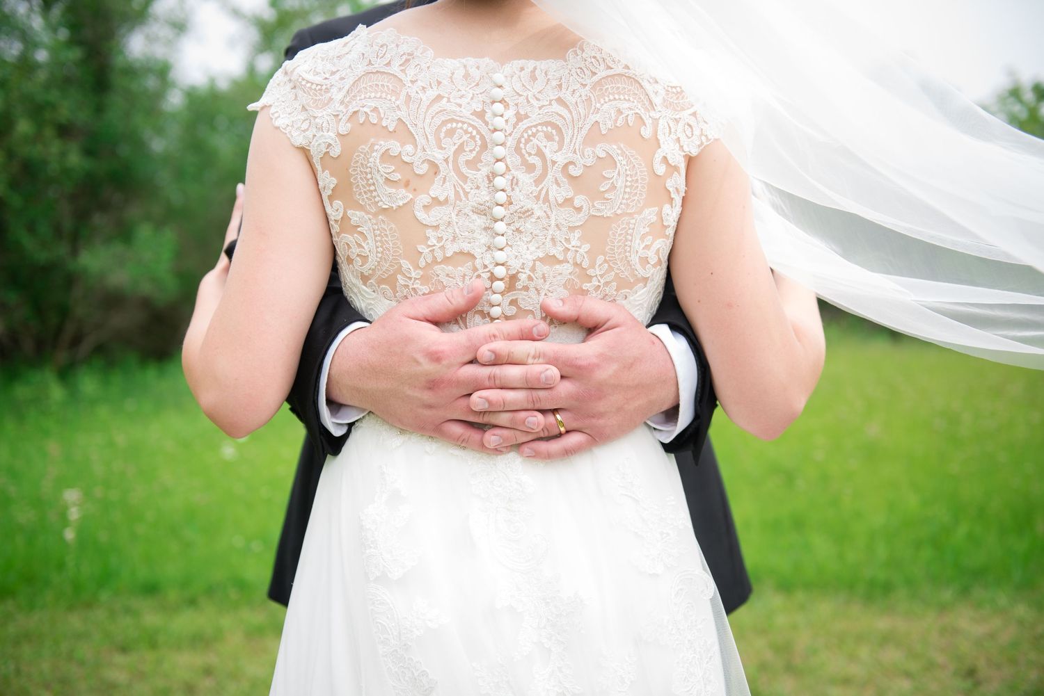 Groom embracing bride from behind, highlighting beautiful lace and pearl button detail on wedding gown.
