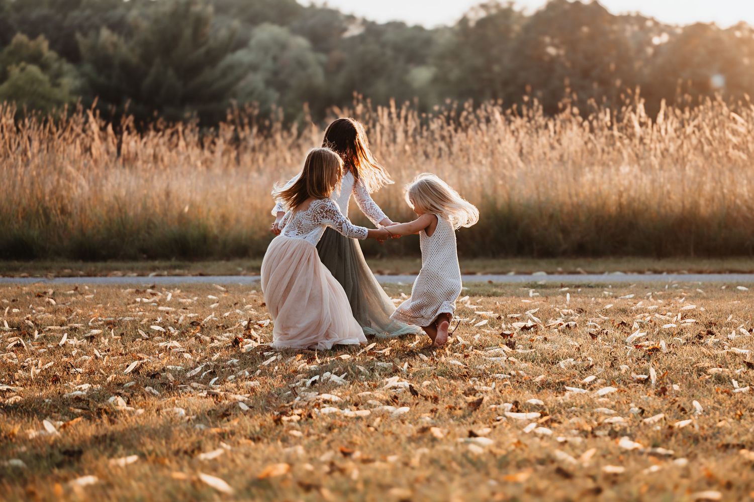 Three girls in white dresses dance and twirl together in an autumn field at golden hour.