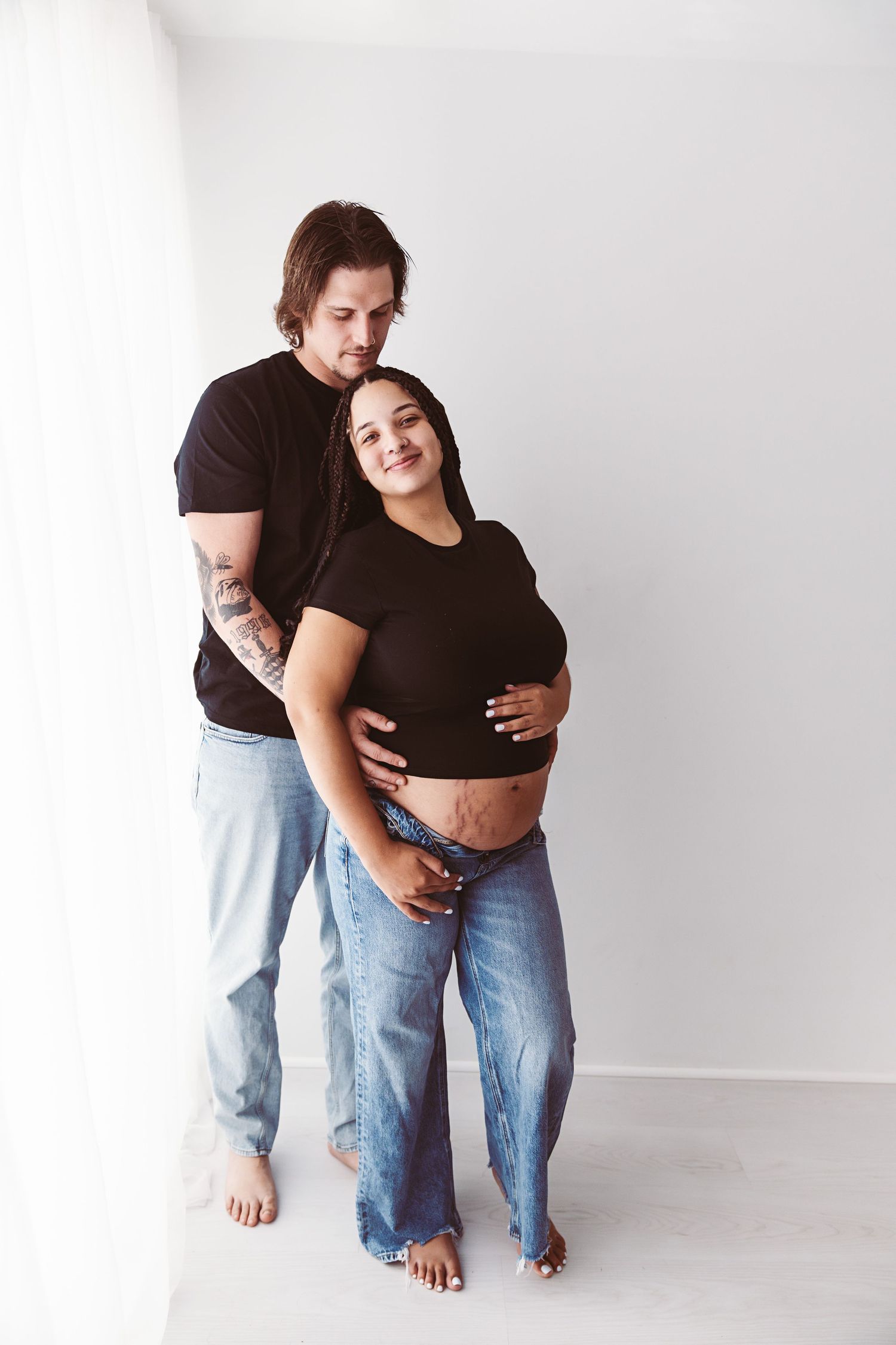 Couple in matching black tops and jeans embracing, man holding pregnant partners baby bump.