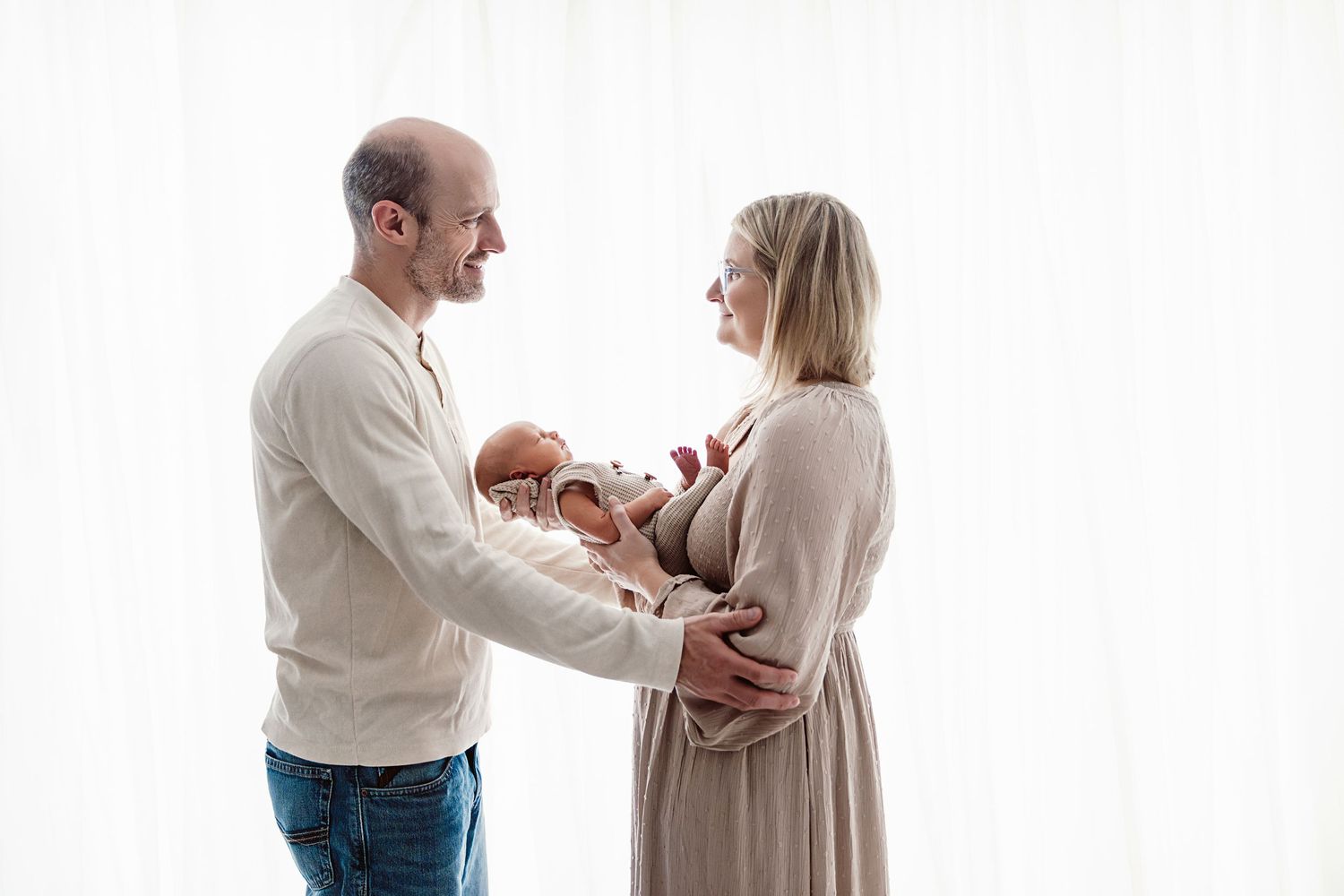 Happy parents in neutral tones admire their newborn baby together indoors.