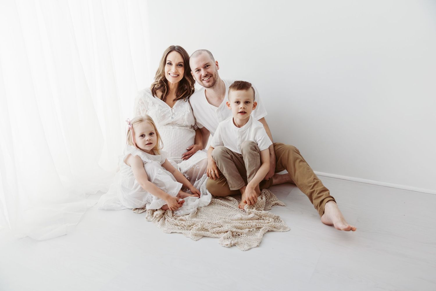 Happy family of four sitting on white floor together in a bright maternity photo session.