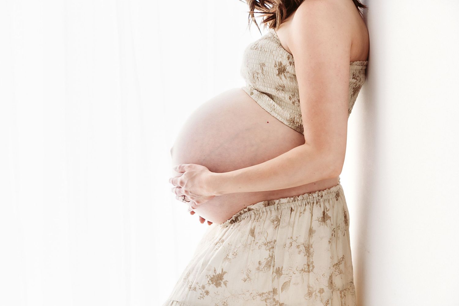 Side profile of pregnant woman in floral dress cradling baby bump against soft white background.