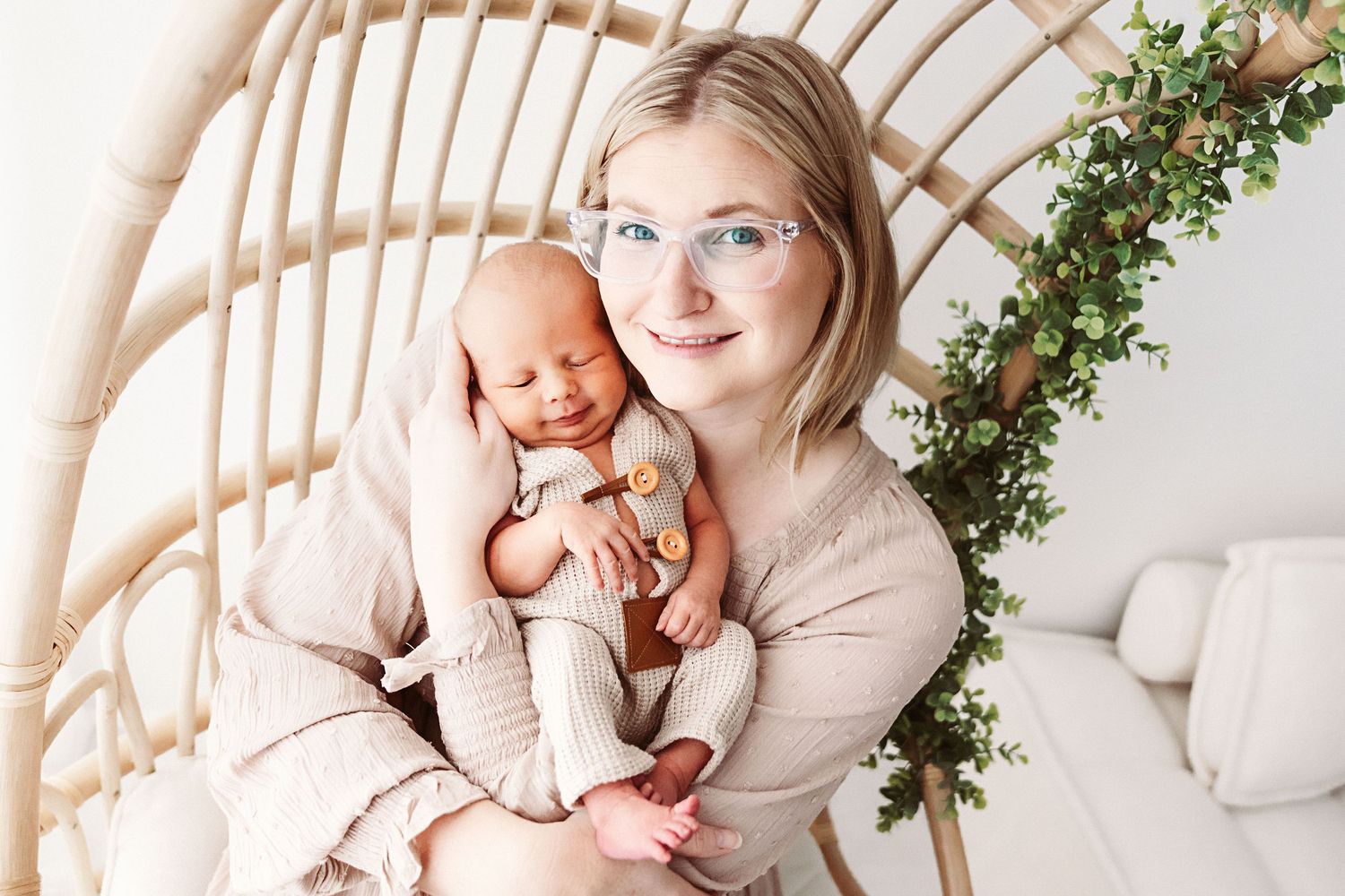 Smiling blonde mother with glasses holds sleeping newborn in boho rattan chair.