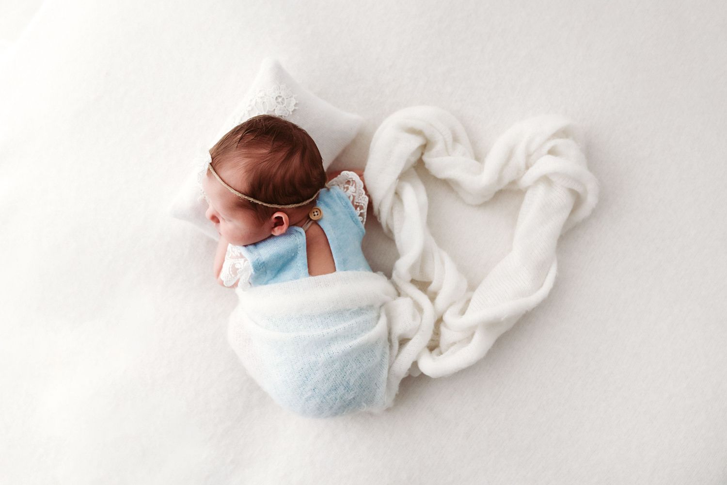 A swaddled newborn baby in blue lies inside a heart shape made from white fabric on a white background.