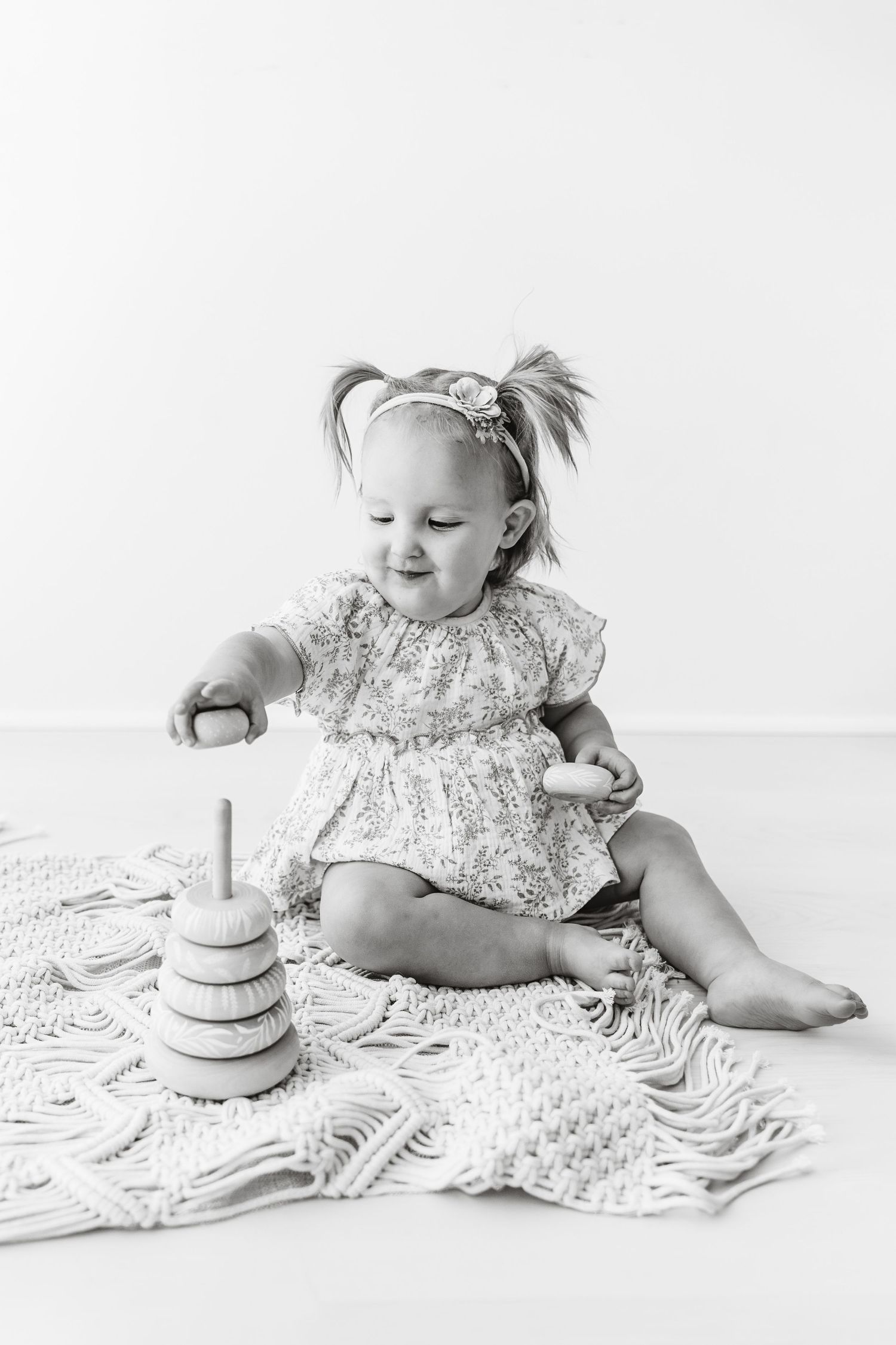 Black and white photo of a toddler girl playing with a wooden stacking toy on a macrame blanket.