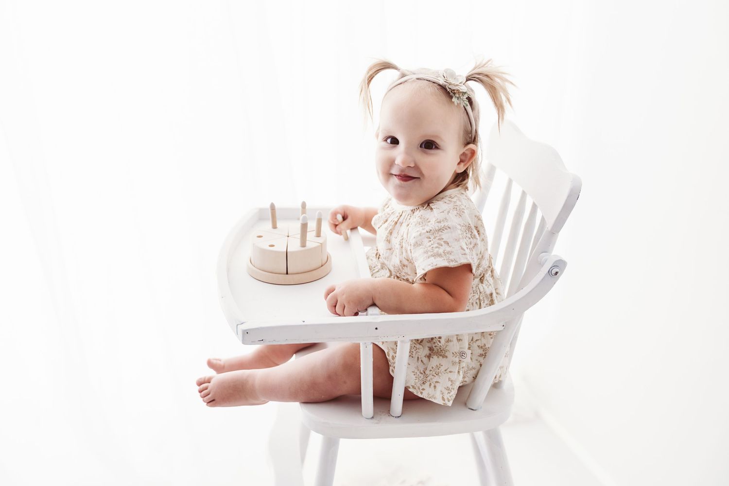 Smiling toddler girl with pigtails sitting in a white high chair with a wooden birthday cake toy.