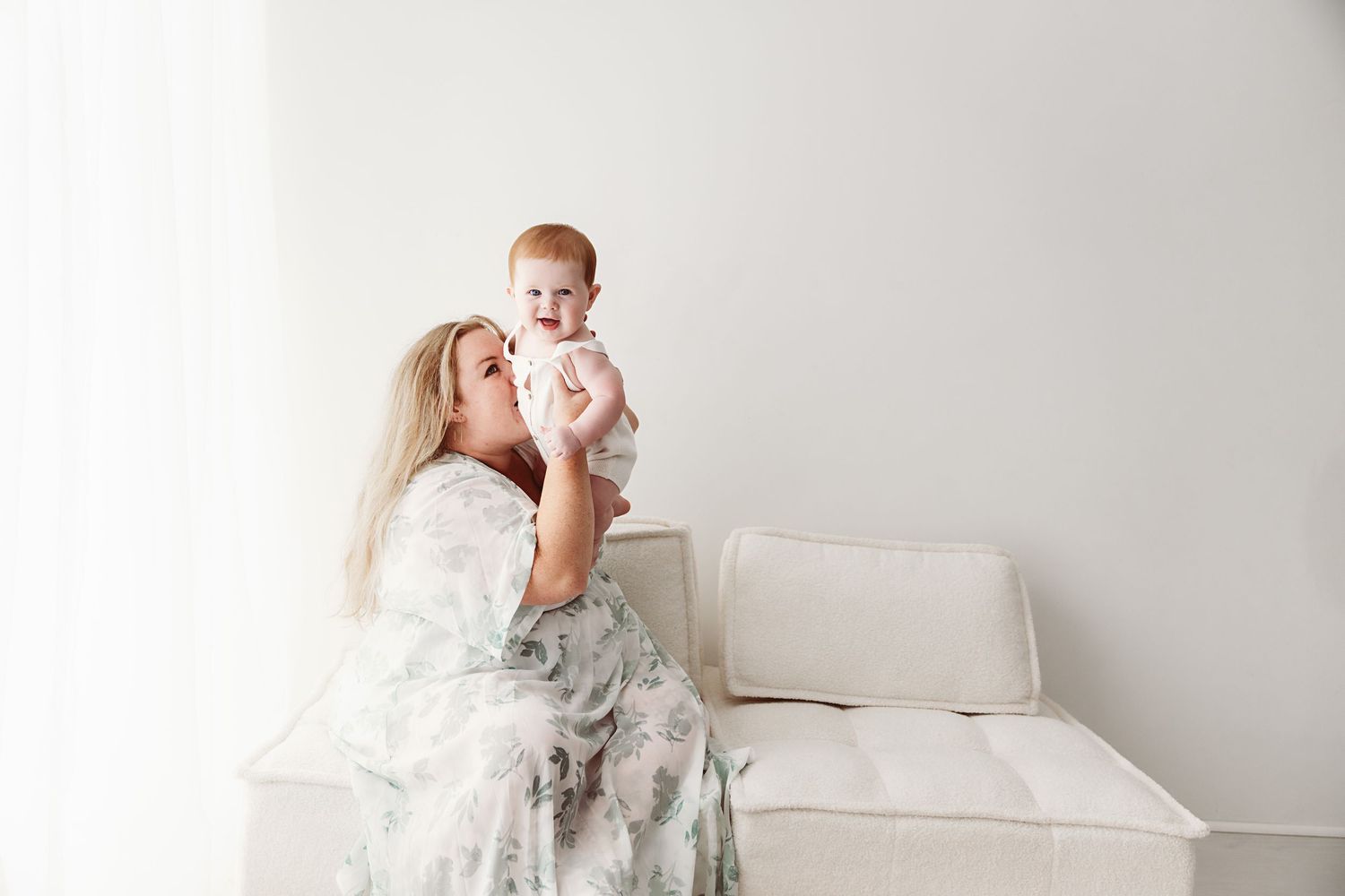 A blonde mother joyfully lifts her smiling baby girl up in a bright white room.