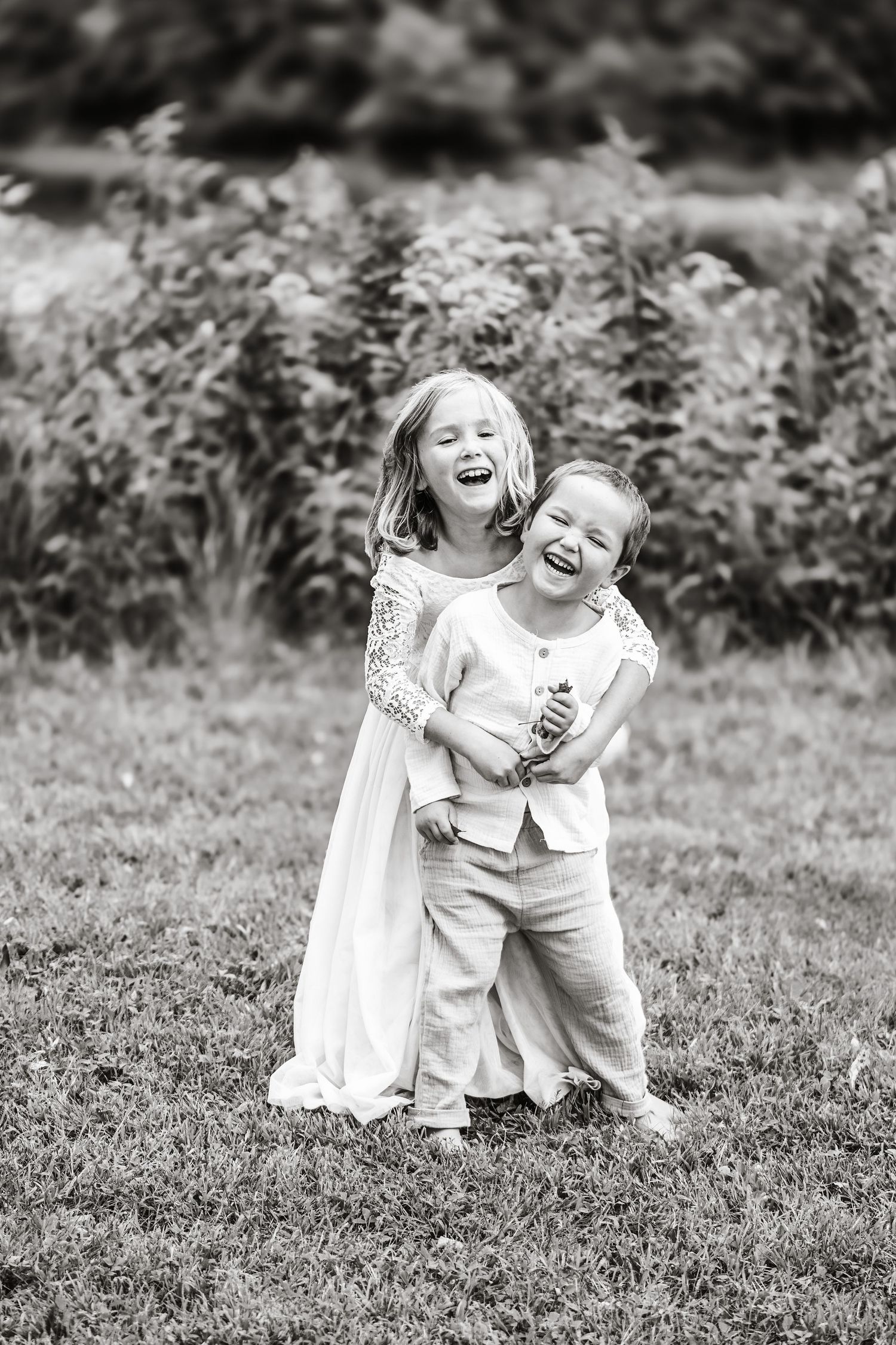 Black and white photo of two laughing children hugging in a garden setting.